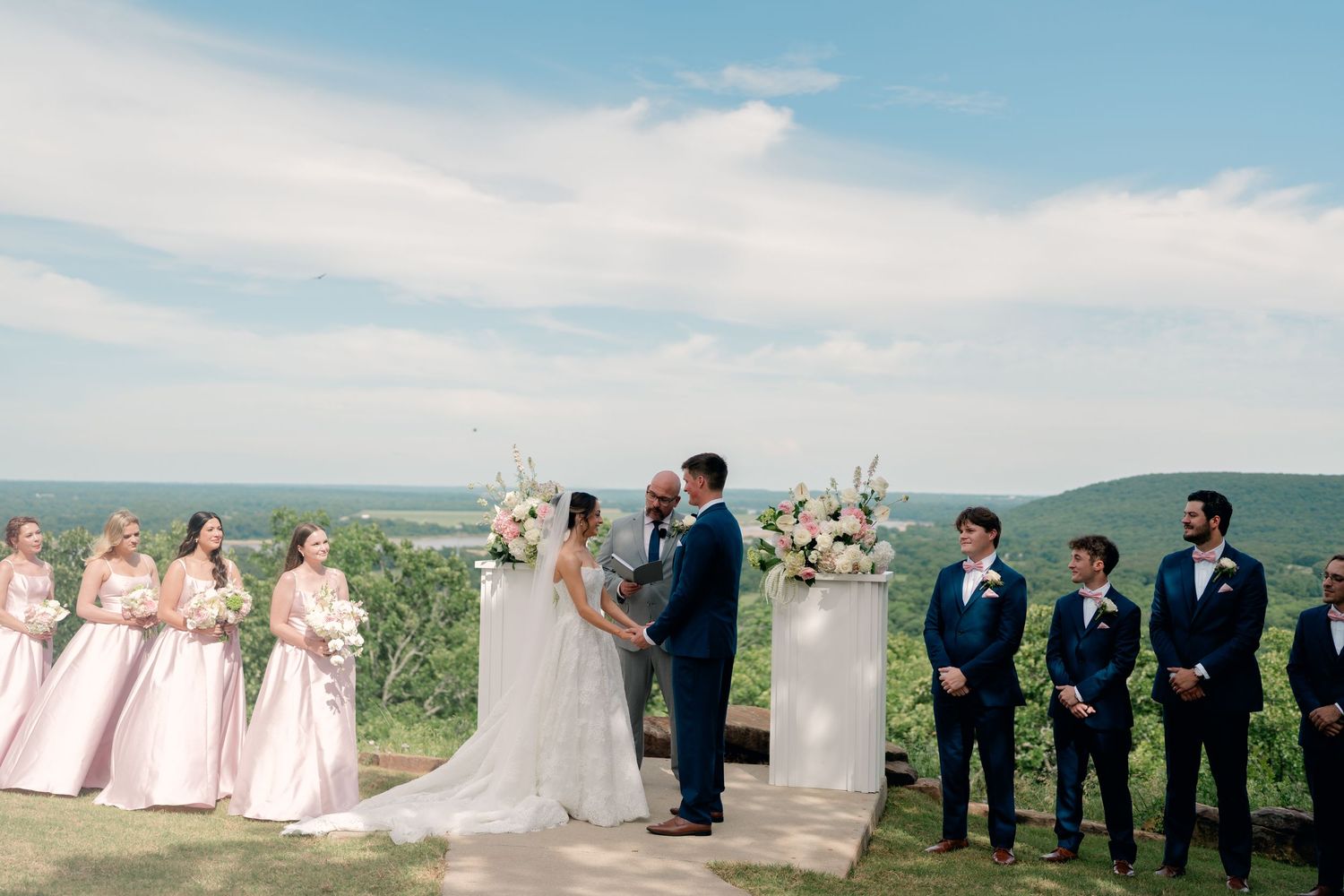 Outdoor wedding ceremony with bridesmaids in pink gowns and groomsmen in navy suits, set against a scenic mountain landscape.