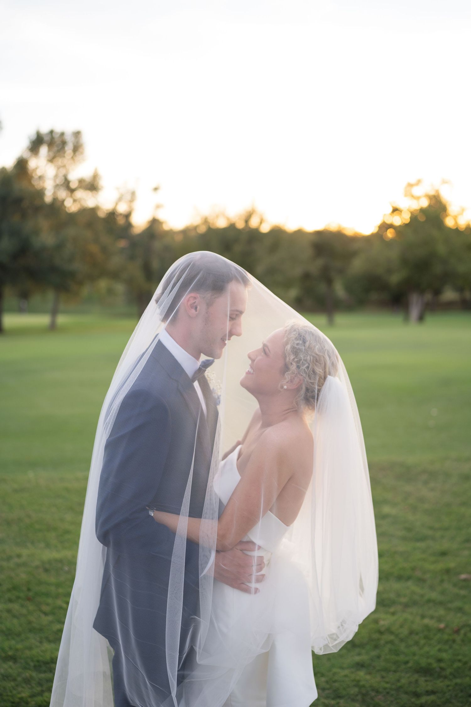 A romantic wedding portrait under a veil during golden hour on a golf course.