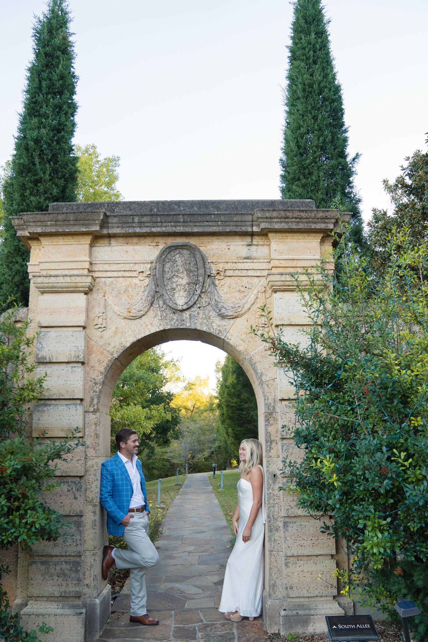A couple poses beneath a decorative stone archway in a formal garden setting.