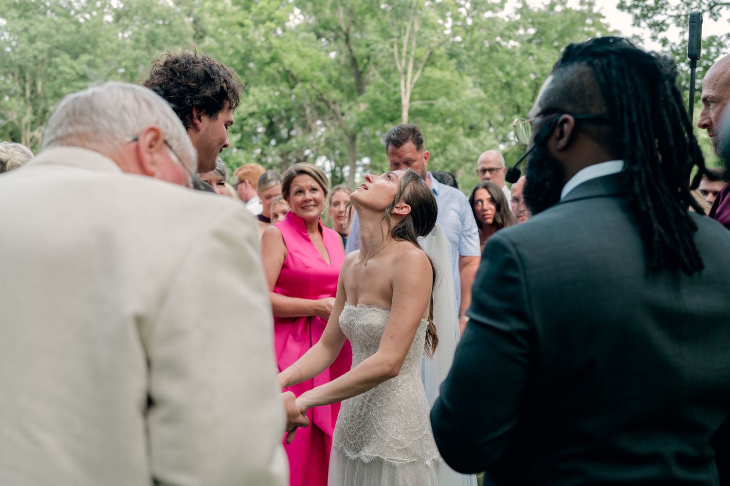 Bride in beaded gown laughing with guests at an outdoor wedding ceremony surrounded by trees.