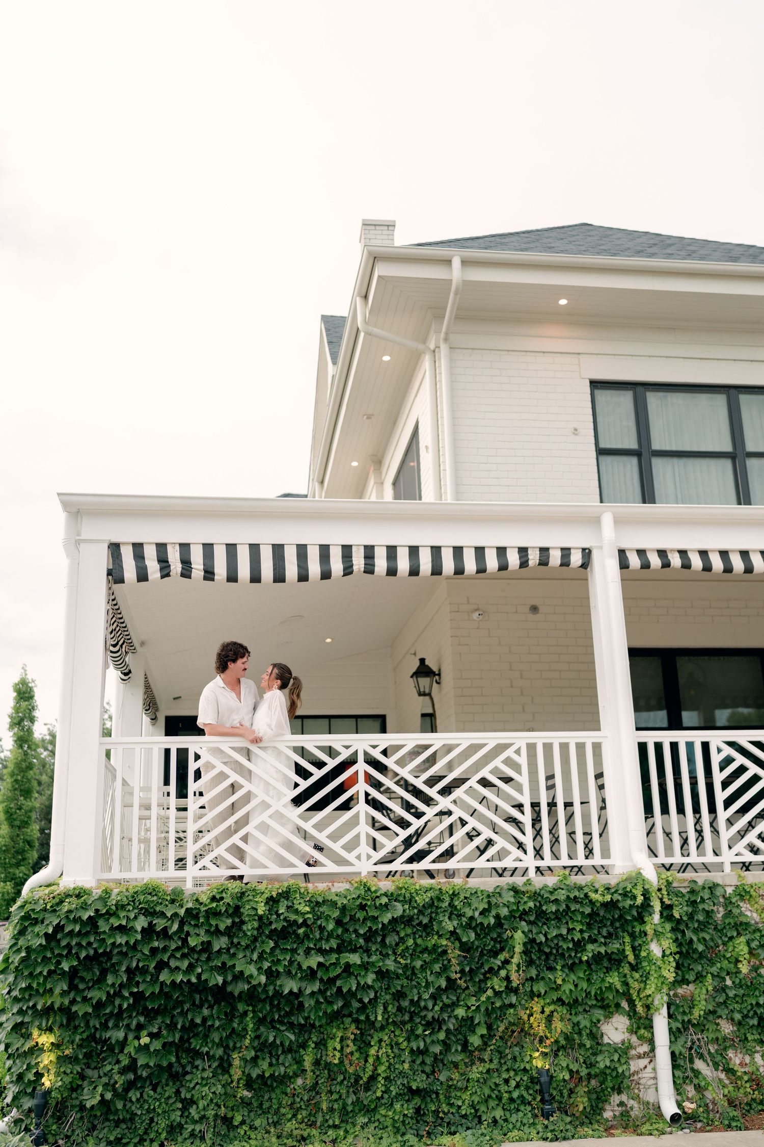 Newlywed couple sharing a romantic moment on a white porch with striped awning surrounded by lush green ivy.