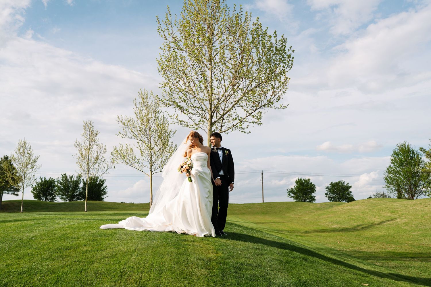 Bride and groom walk hand-in-hand on lush green golf course with blooming trees under blue sky.