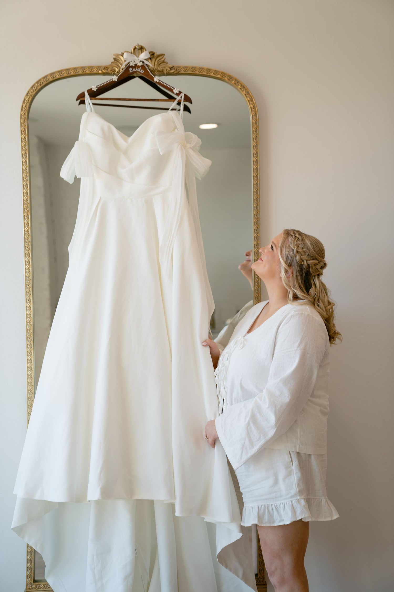 White wedding dress hanging on display next to ornate gold mirrors.