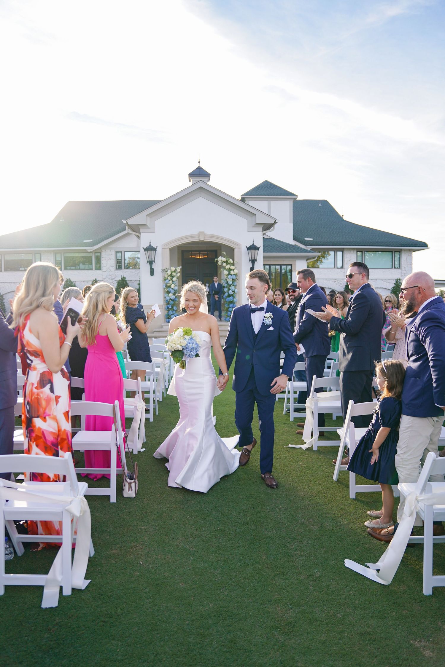 Wedding guests gather on white chairs during an outdoor ceremony at sunset.