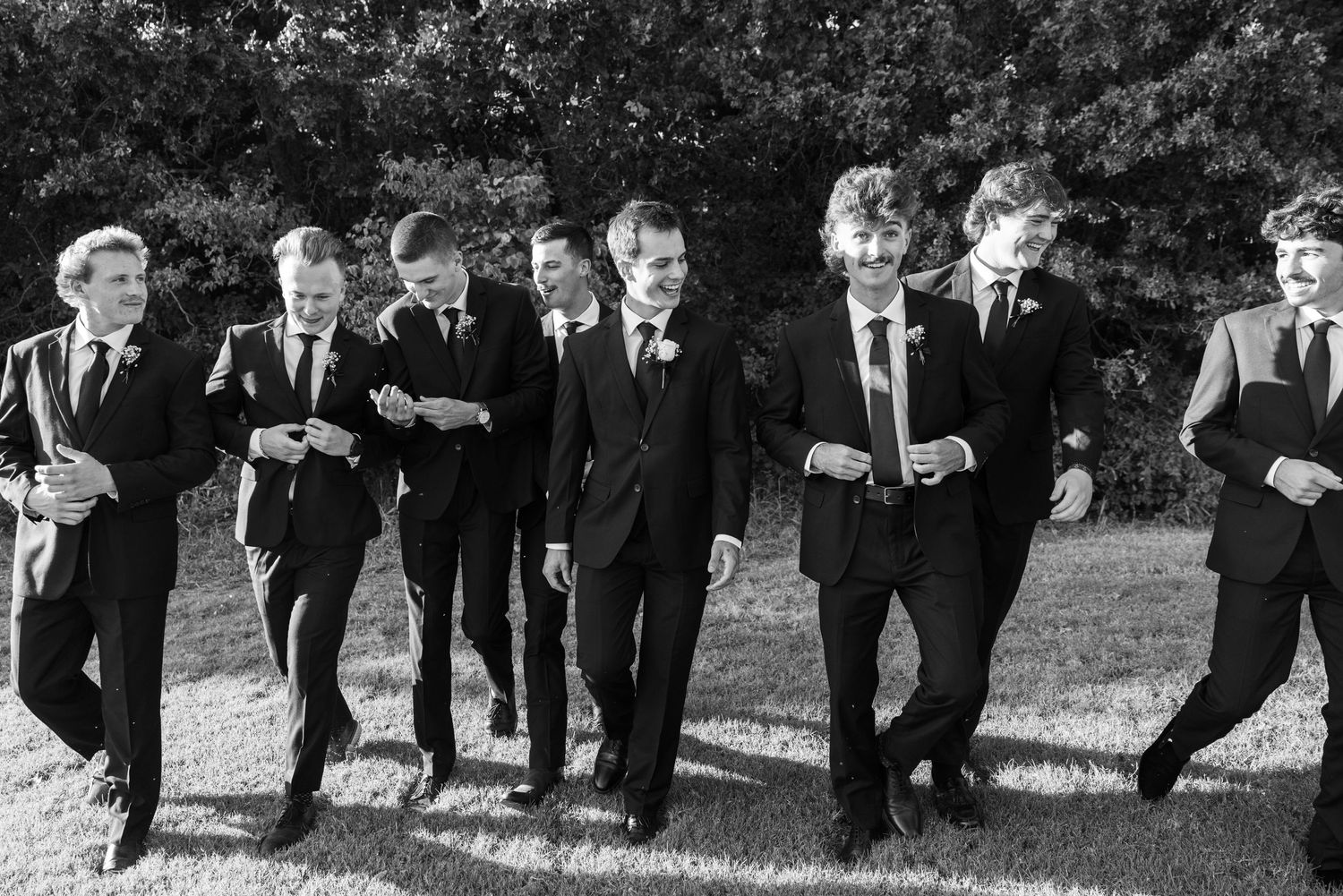 Groomsmen in black suits walking in formation during an outdoor wedding photoshoot.
