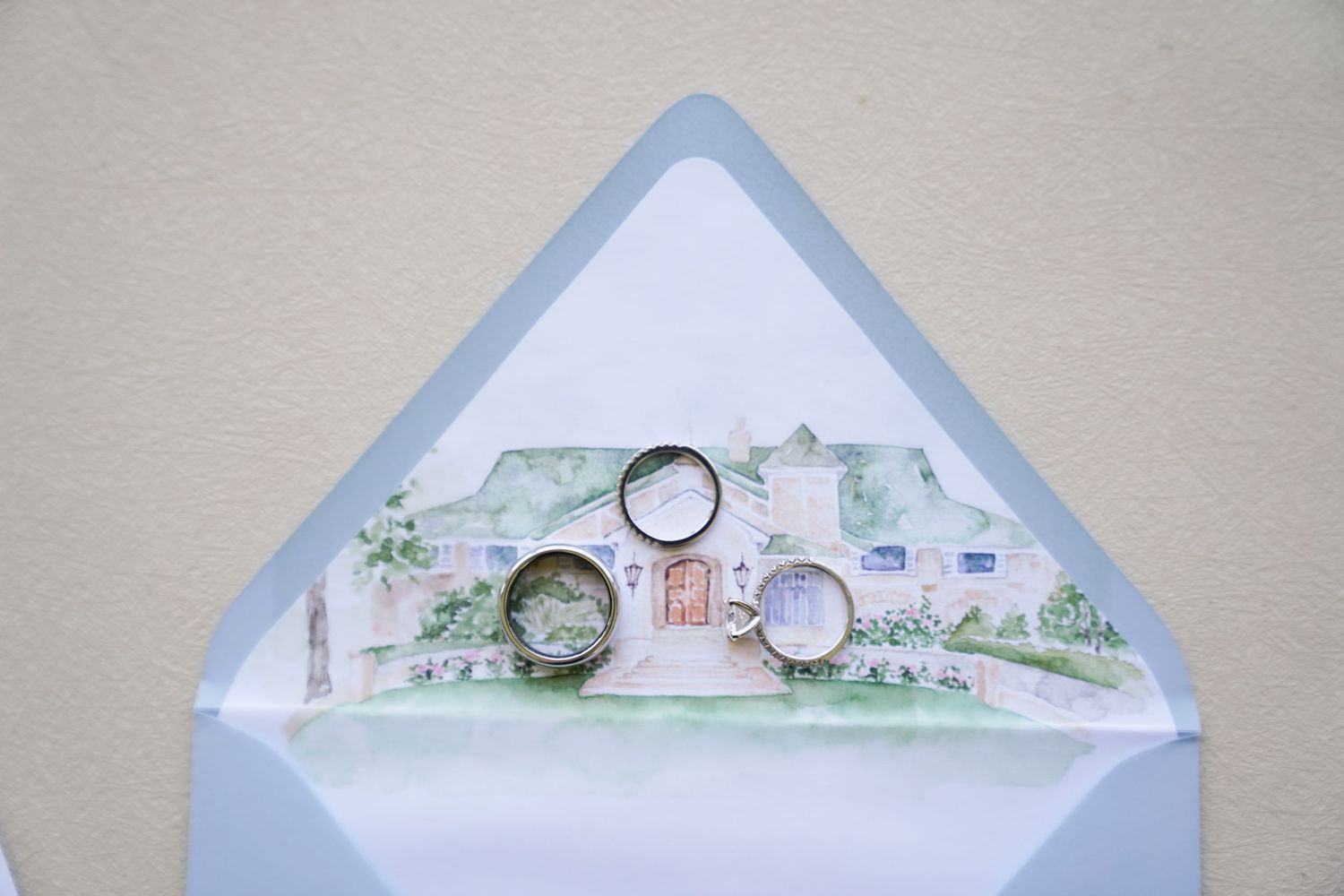 Wedding rings displayed on light blue envelopes with watercolor house illustrations.