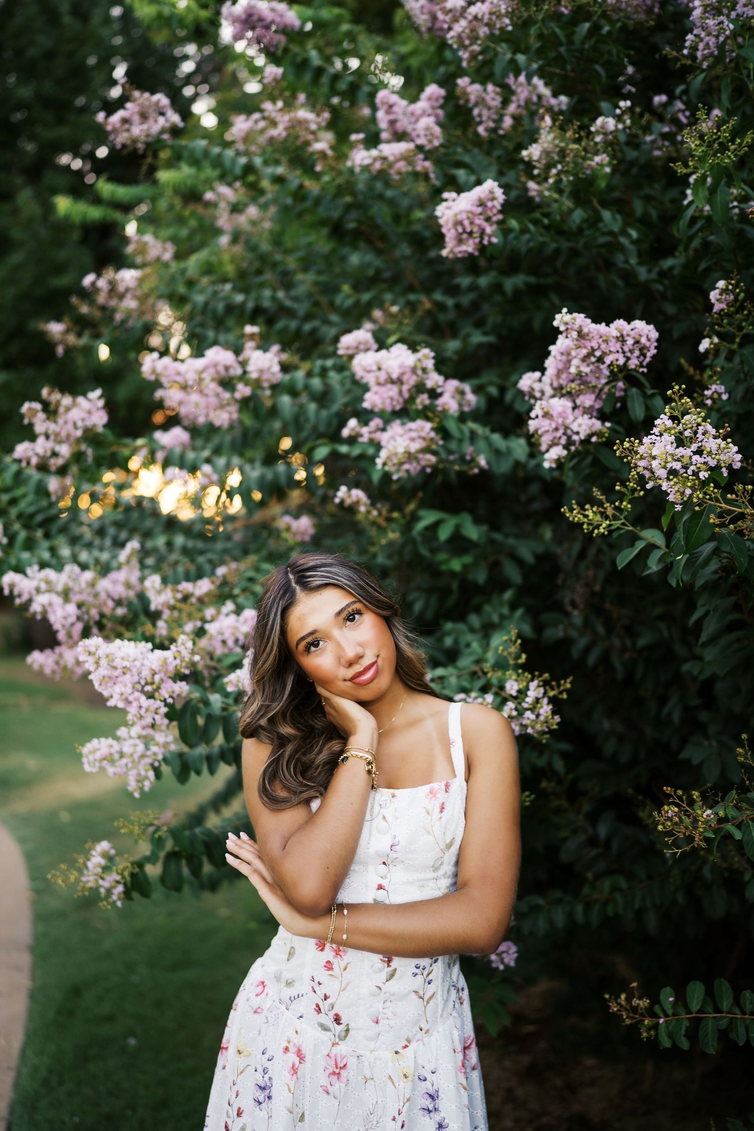 A white floral dress is worn in a garden setting with blooming pink lilac bushes on a summer evening.