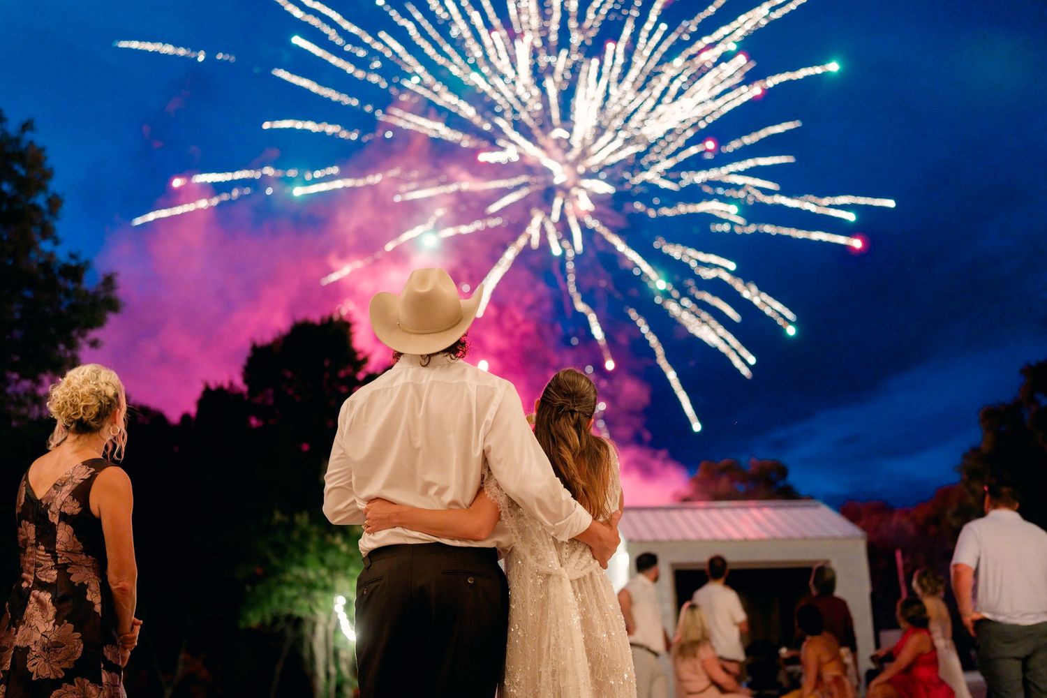 Couple embraces while watching colorful fireworks light up the night sky at outdoor wedding reception.