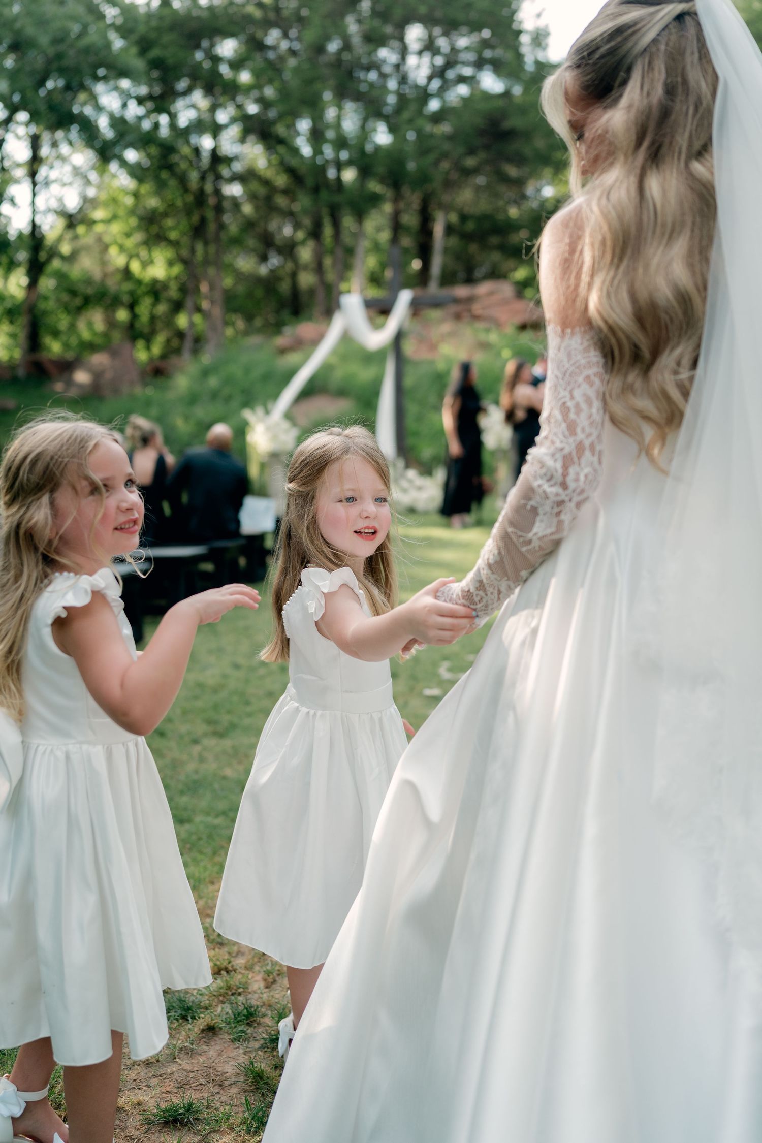 Two flower girls in white dresses hold hands with a bride in a lace gown at an outdoor wedding ceremony.