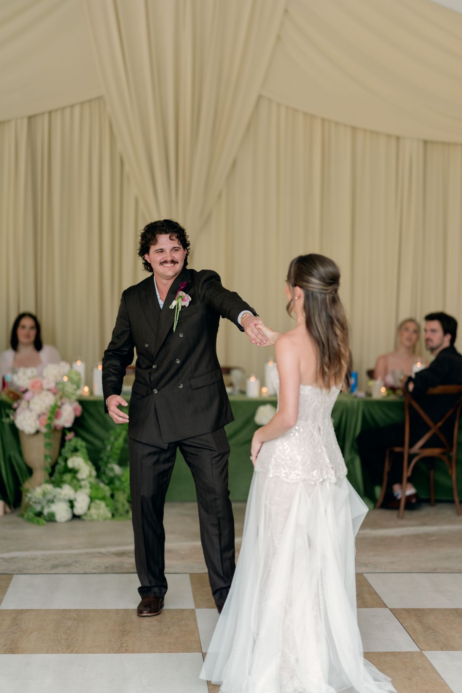 Groom in black suit extends hand to bride in strapless gown for first dance in elegant draped reception tent.