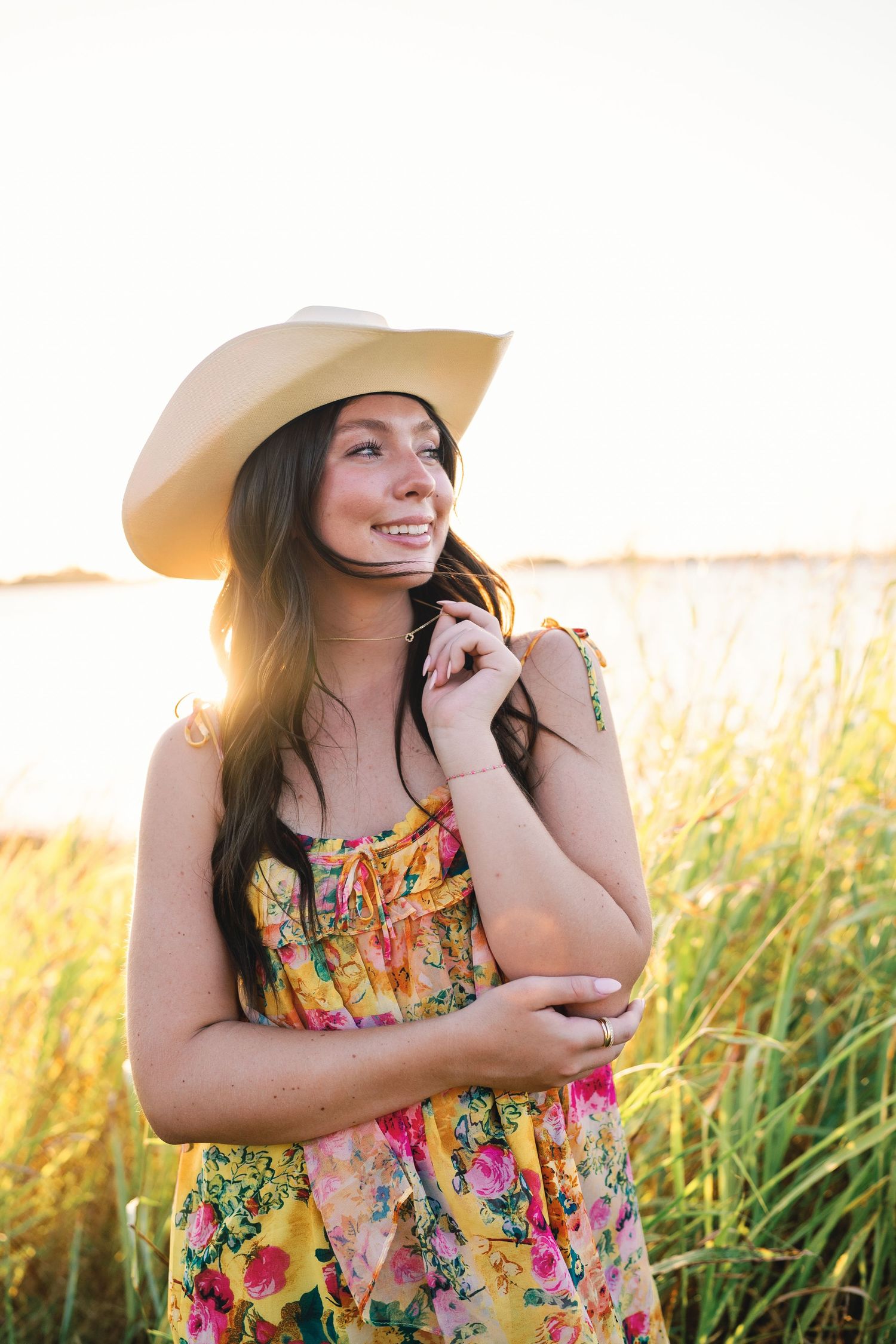 Woman in floral dress and sunhat poses in golden sunlit field during sunset.