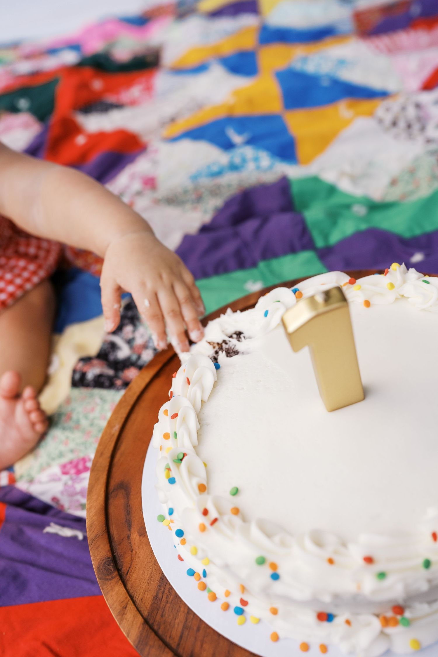Close-up of hands decorating a white cake with colorful sprinkles on a patchwork quilt background.