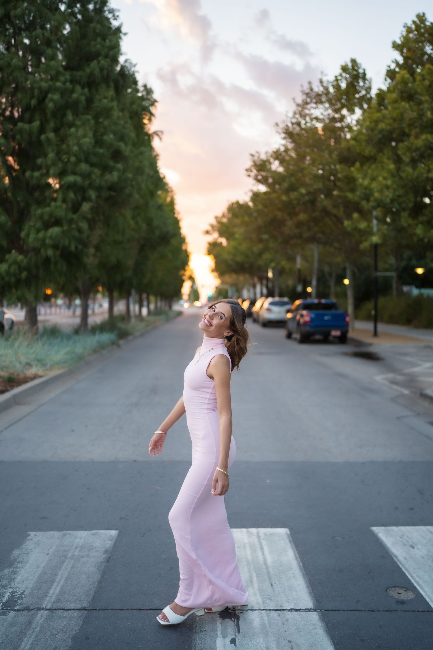 A person in a pink dress dances playfully across a crosswalk at sunset.