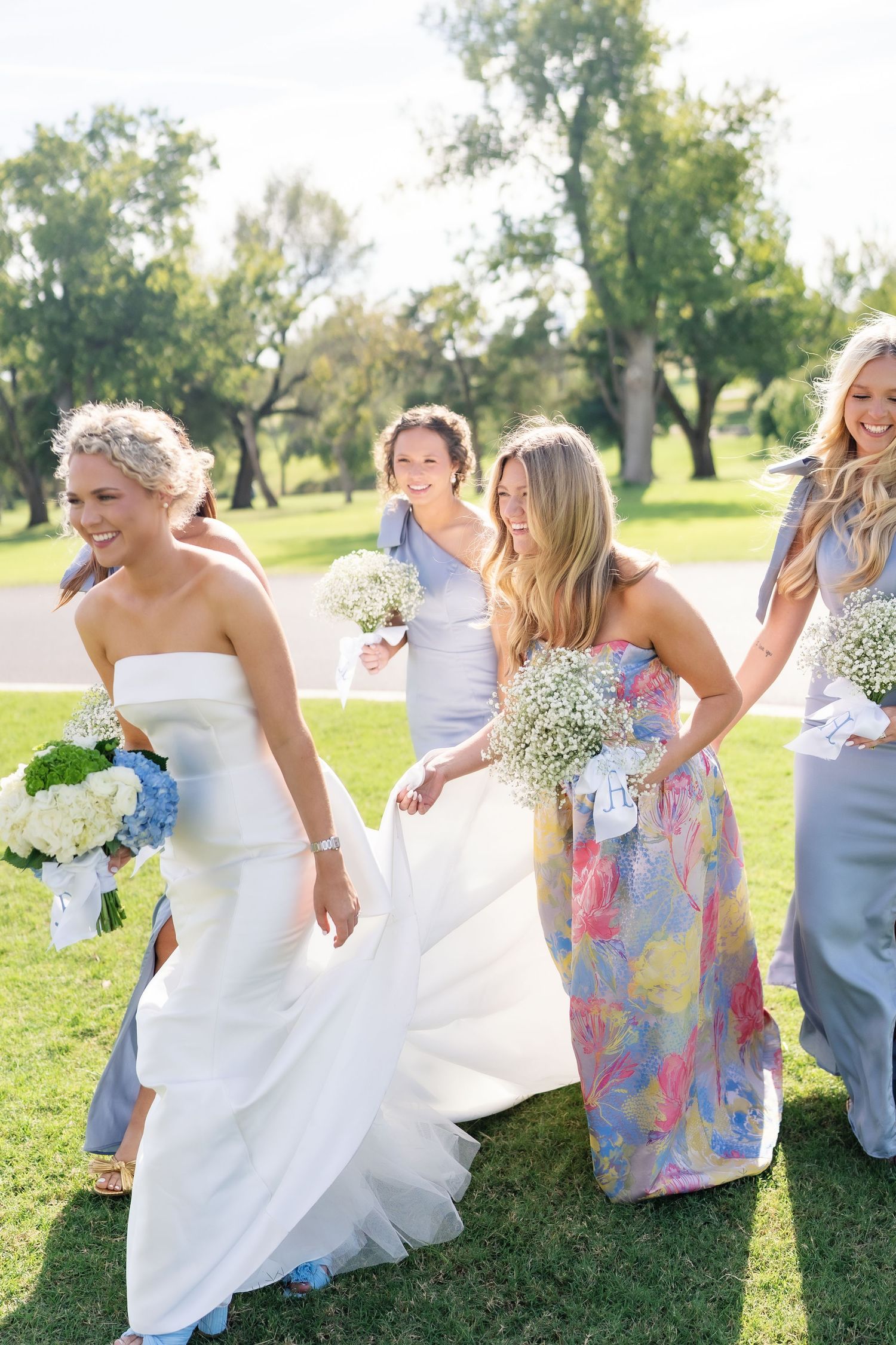 A bridal party in colorful dresses laughs and dances together on a sunny lawn.