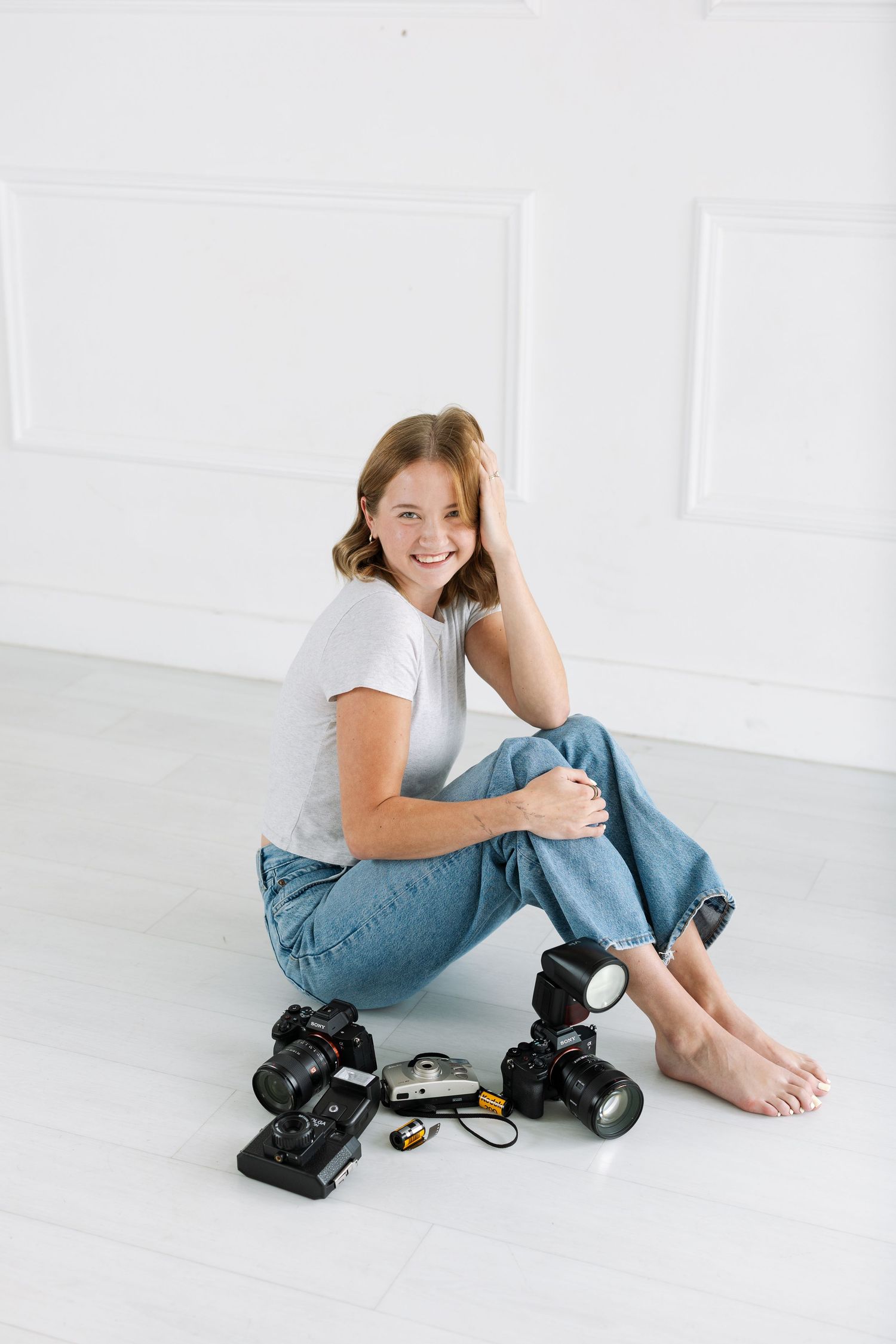 Smiling woman in white tee and jeans sits barefoot on studio floor surrounded by professional cameras and photography equipment.