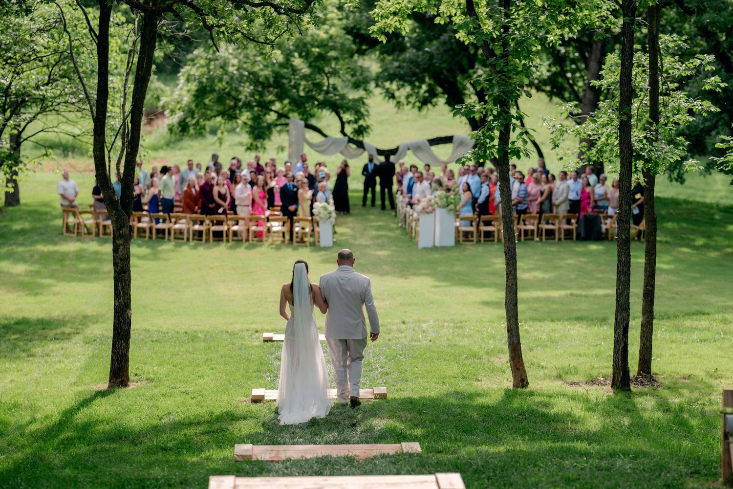 Bride and groom walk hand-in-hand toward outdoor wedding ceremony with guests seated on wooden chairs in lush green park.