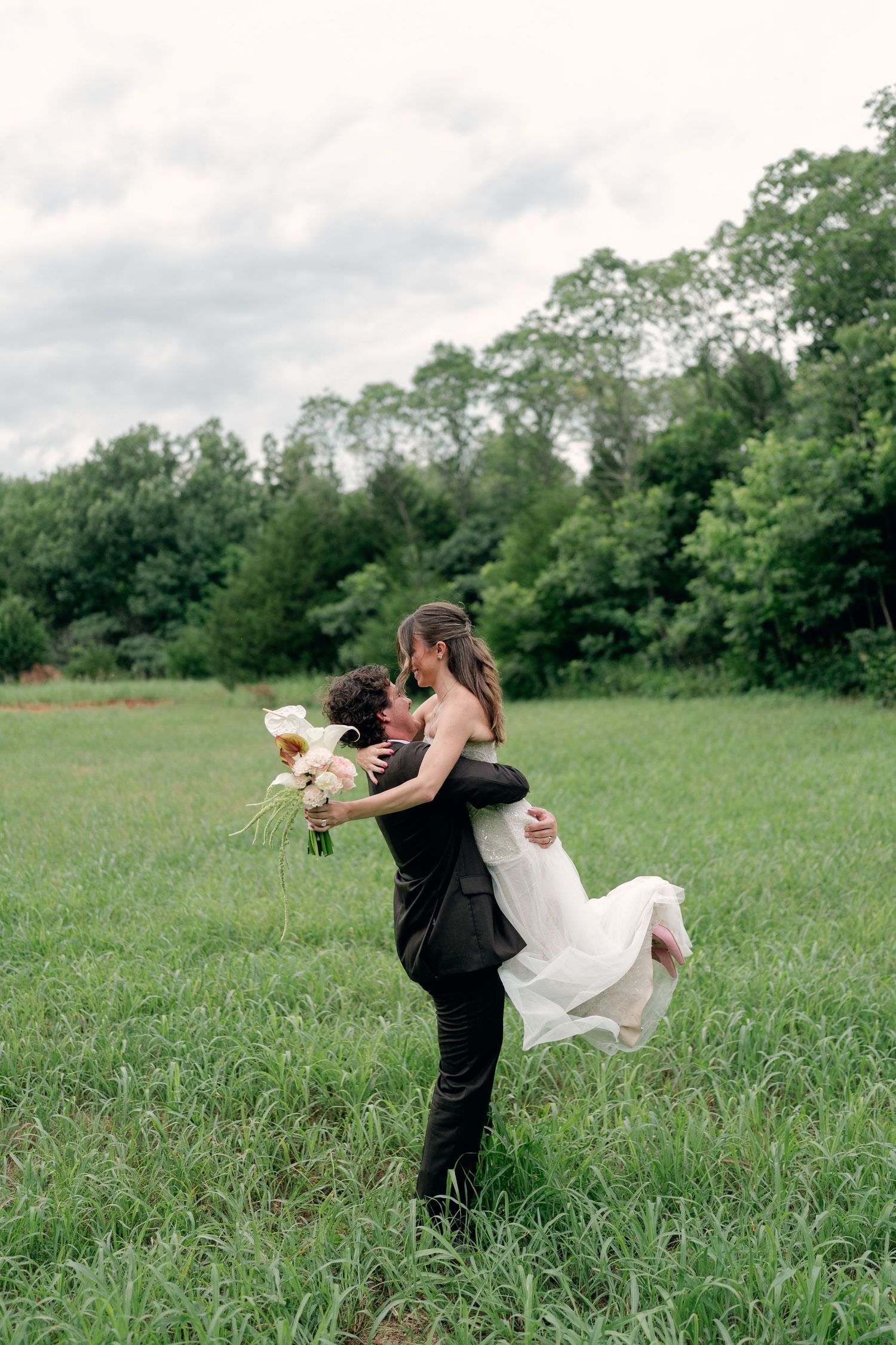 Groom lifts bride in white dress holding bouquet in lush green field with trees backdrop on wedding day.