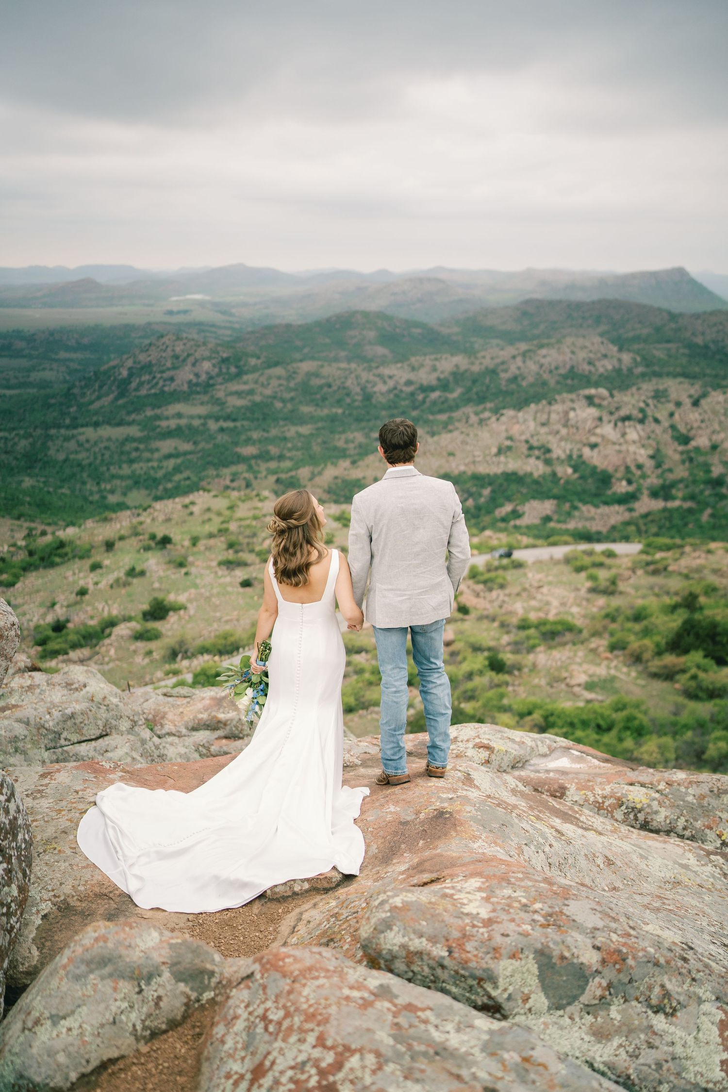 A couple sits on a rocky cliff overlooking a vast mountainous landscape during a photo session.