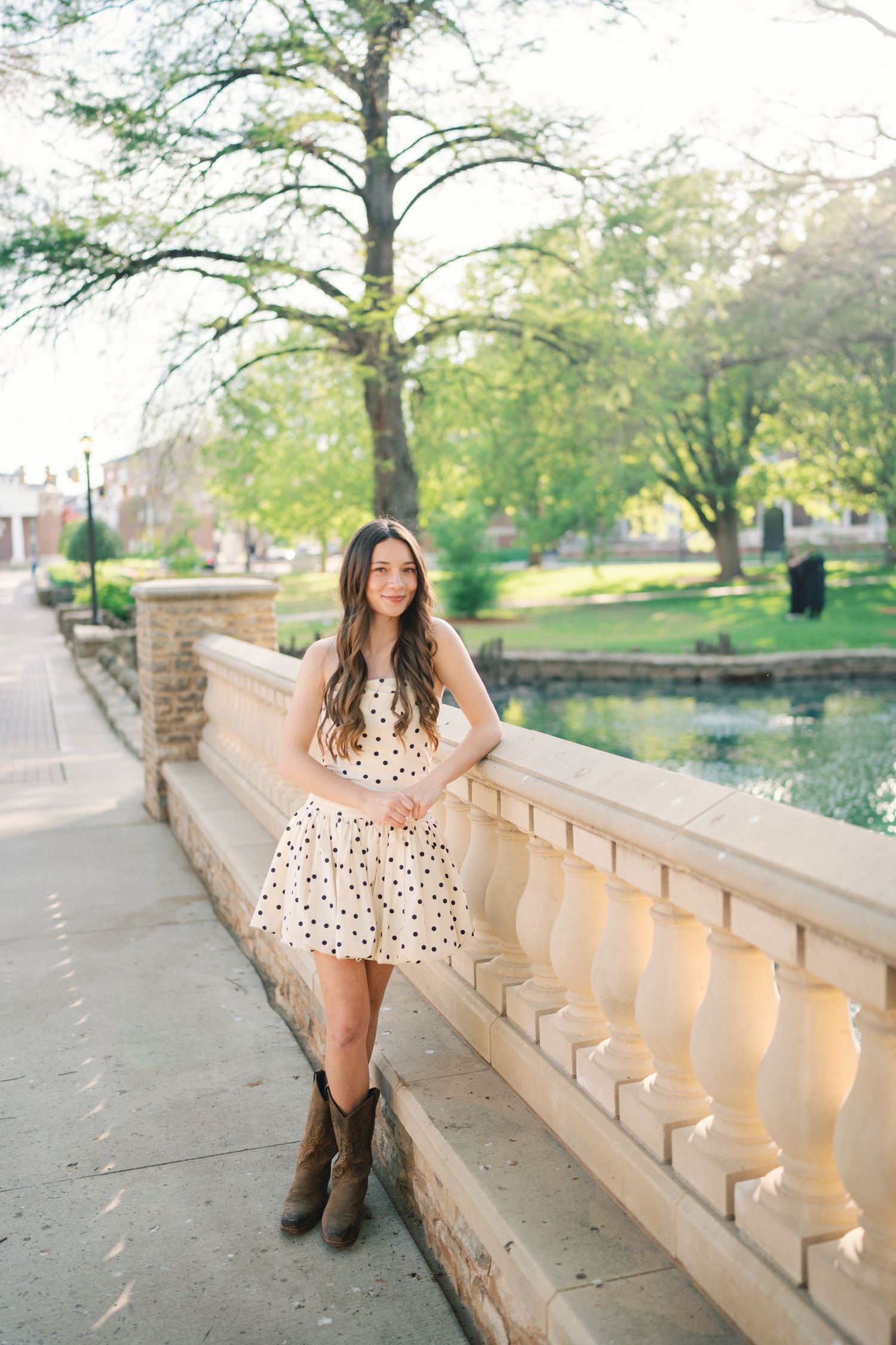 Person in polka dot dress poses along stone railing near pond in springtime setting.