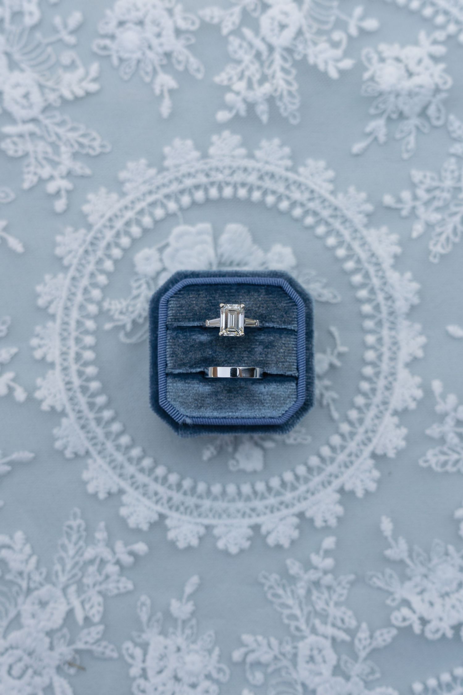 Elegant engagement ring and wedding band in navy velvet box on light blue lace background.