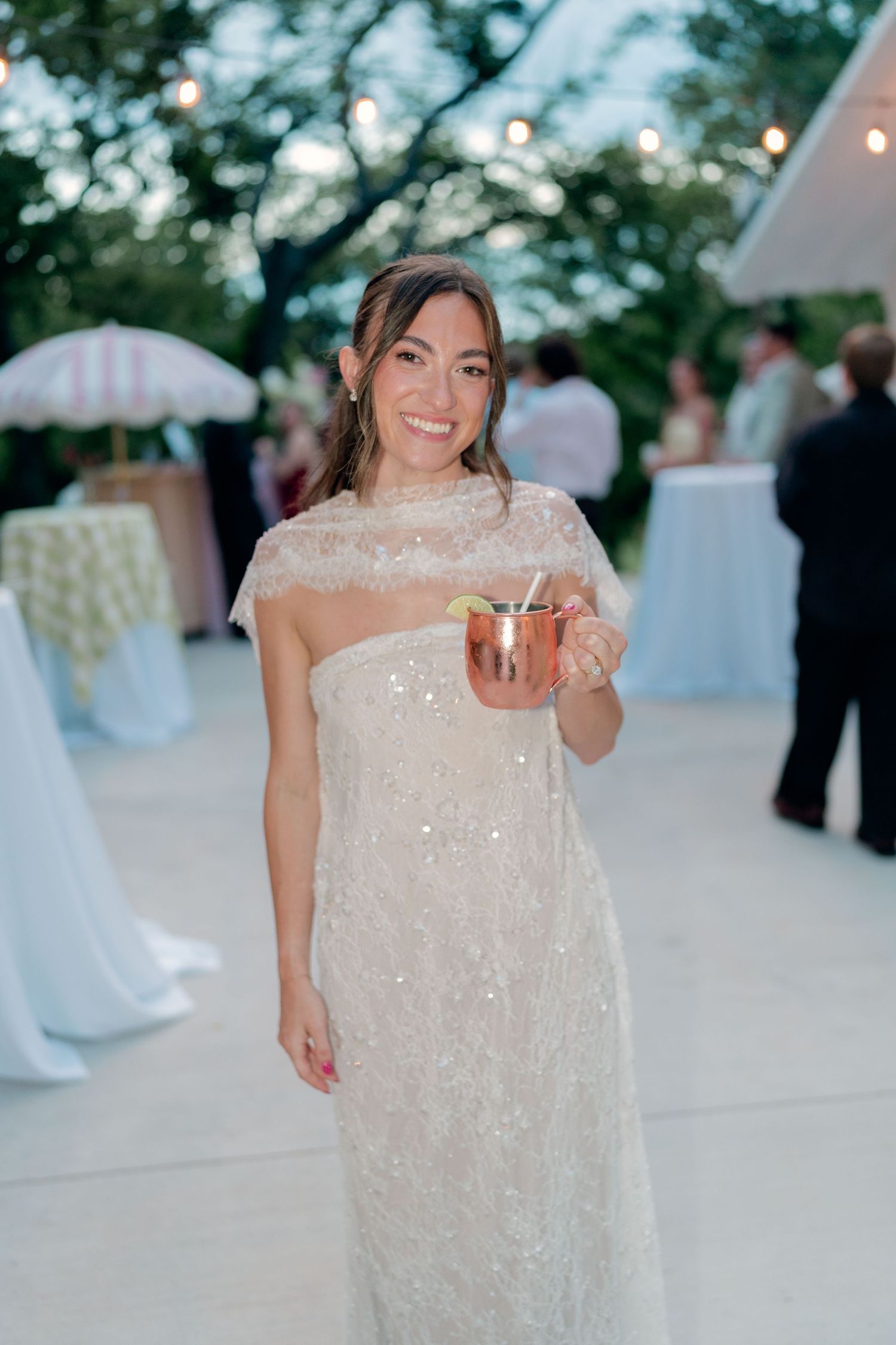 Smiling bride in sequined white gown with cape holding copper Moscow Mule cup at outdoor reception.