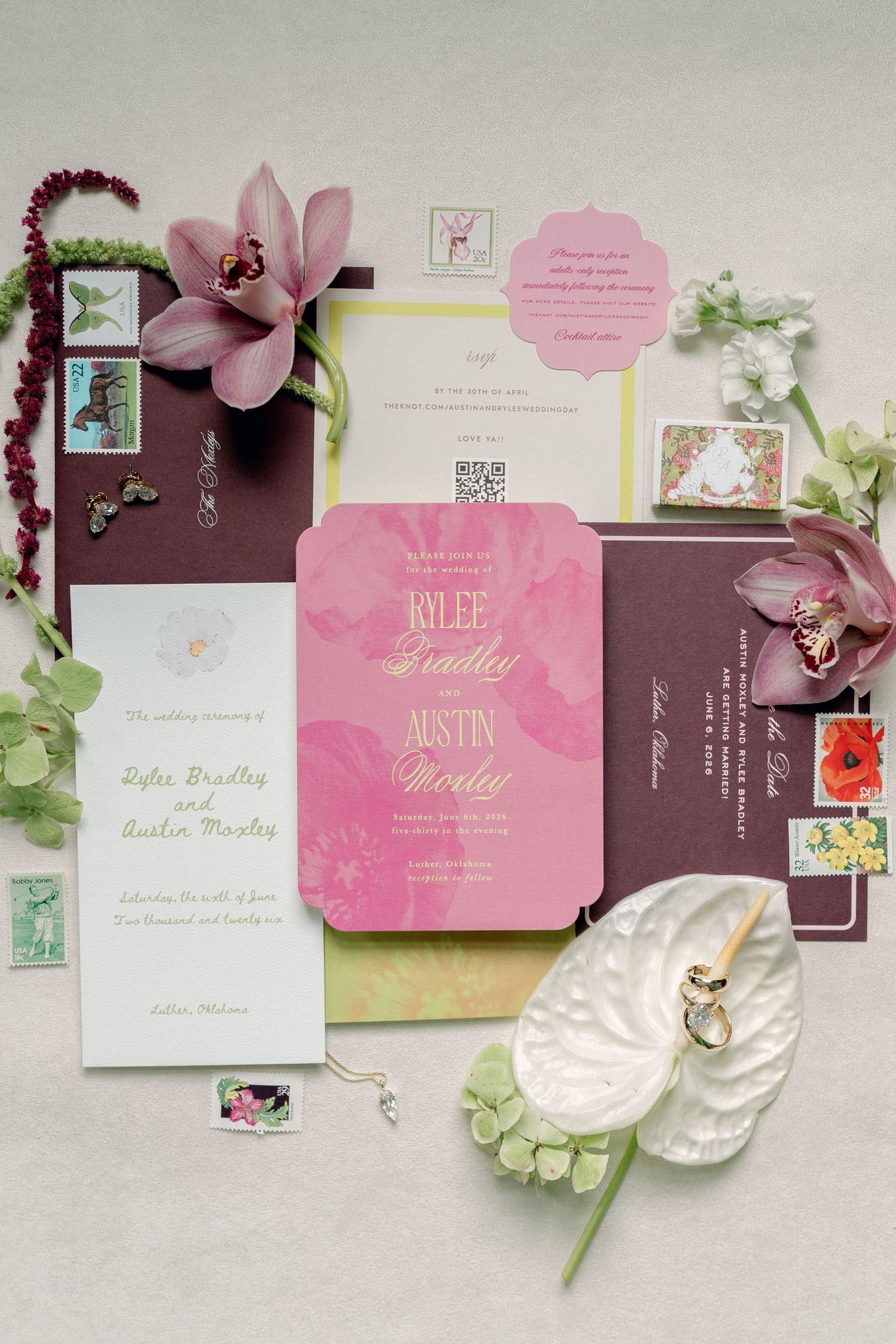 Elegant wedding invitation suite in pink and burgundy with gold script, florals, and decorative stamps on a neutral background.