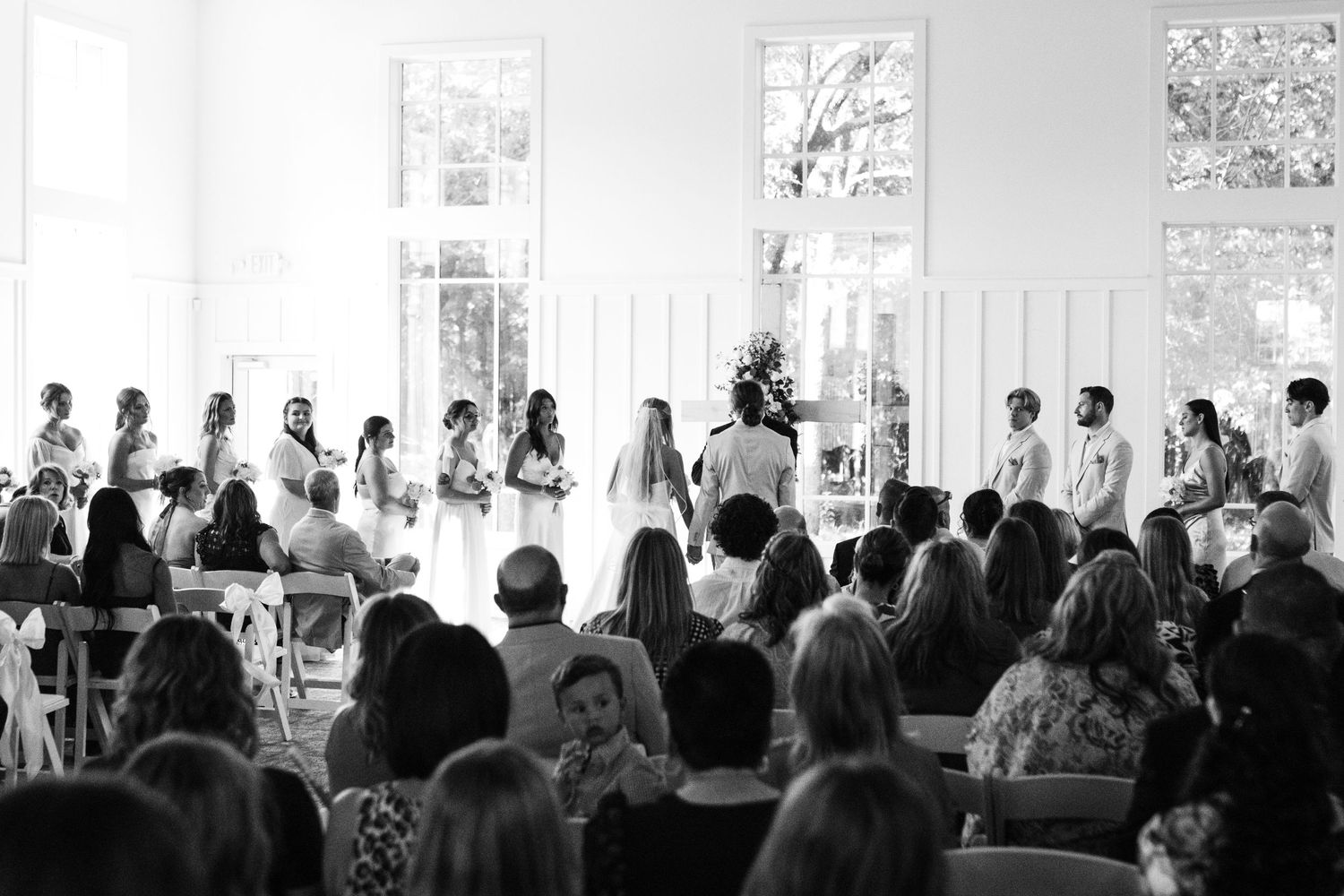 Wedding ceremony in progress inside bright white venue with guests seated.