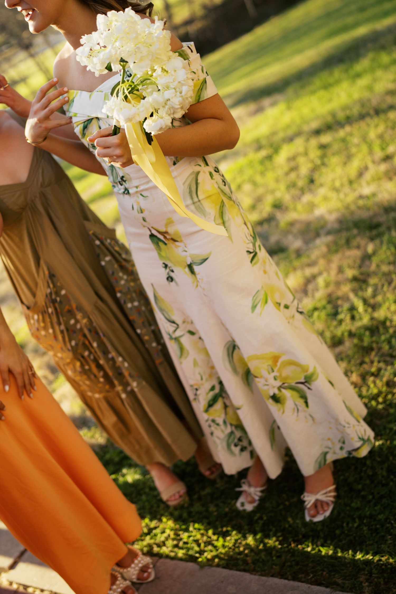 A floral print dress and white bouquet are shown at an outdoor wedding with sunlit grass in the background.