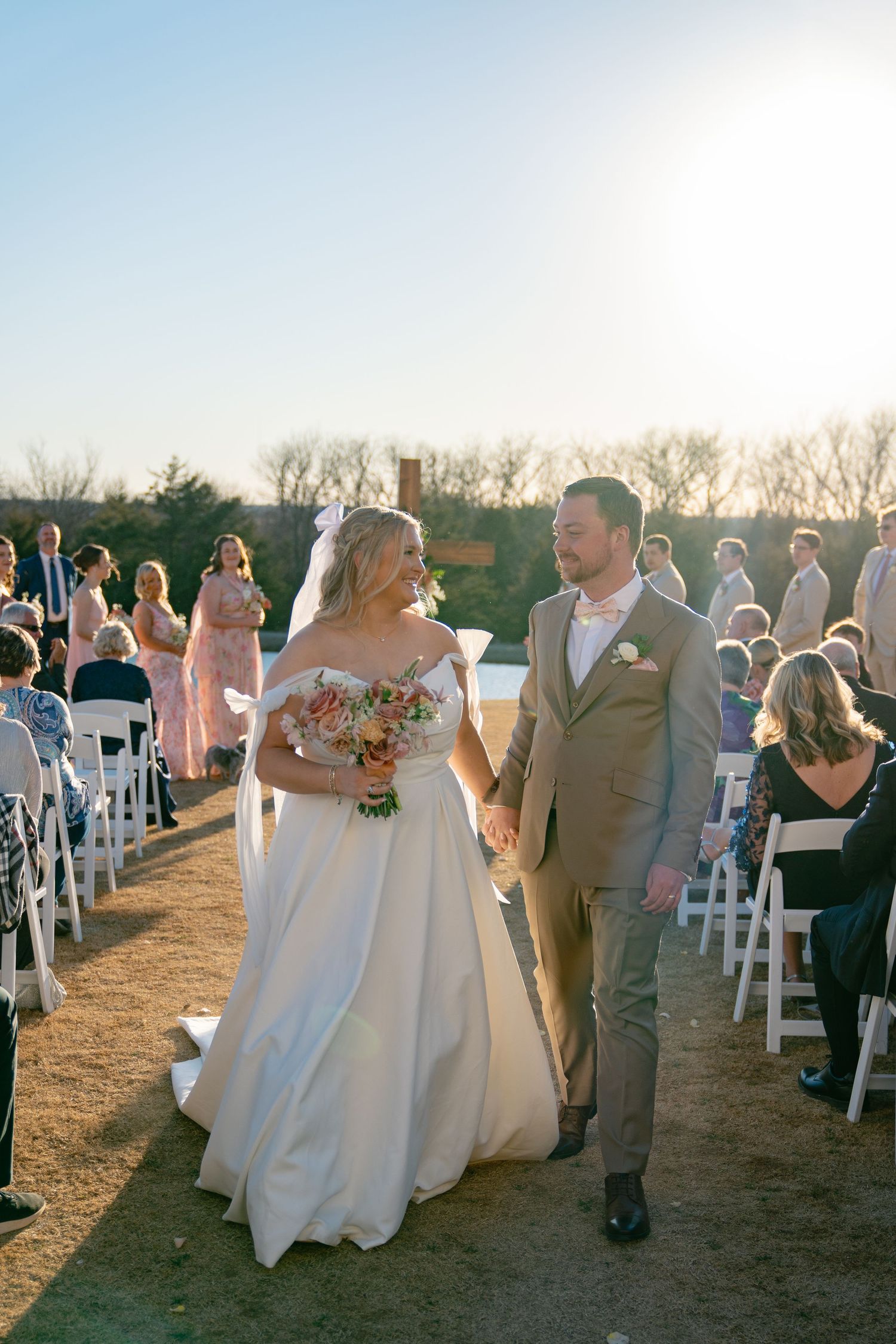 Wedding ceremony procession on outdoor lawn with white chairs and sunset lighting.