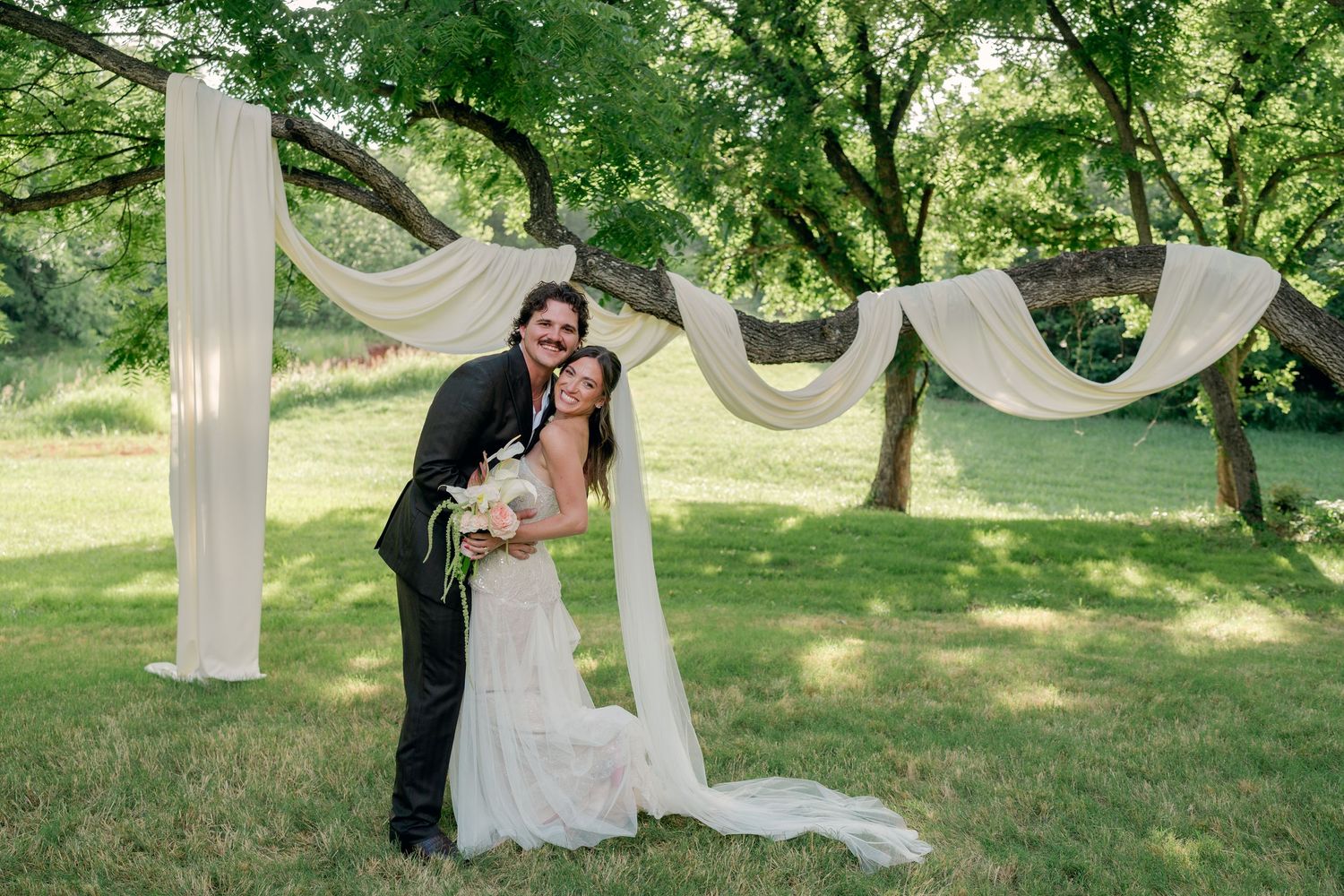 Bride and groom embracing outdoors under white draped fabric wedding arch between lush green trees.