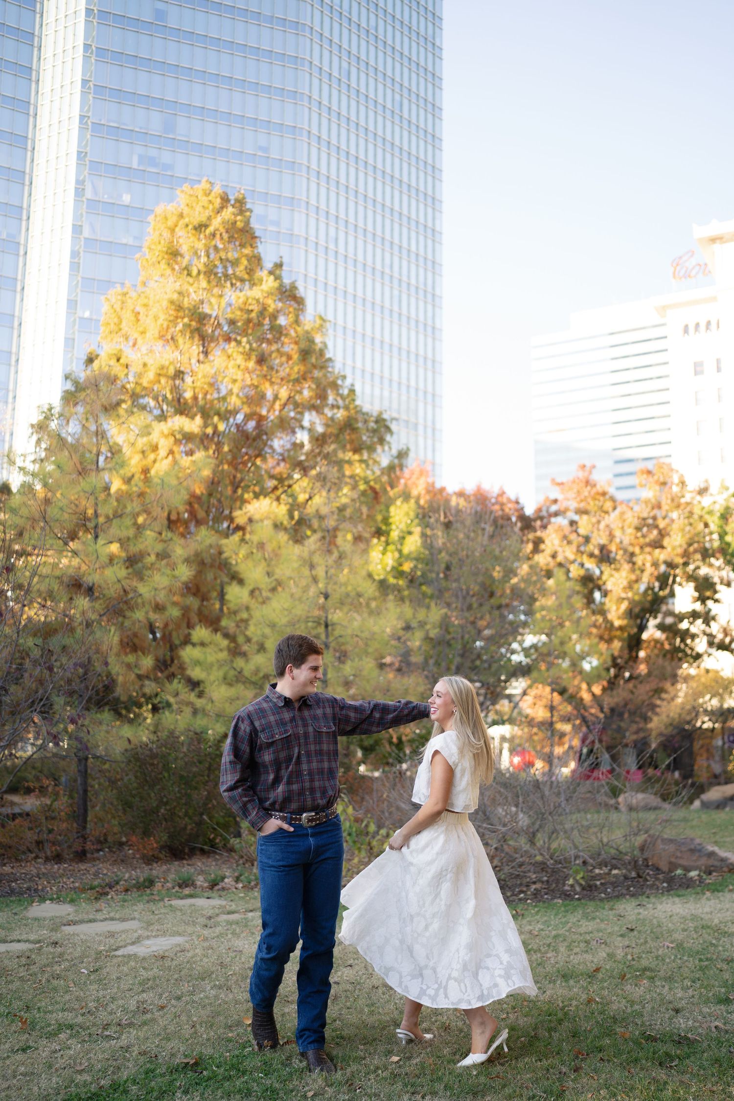 A couple dances playfully in front of a modern glass building during fall season.