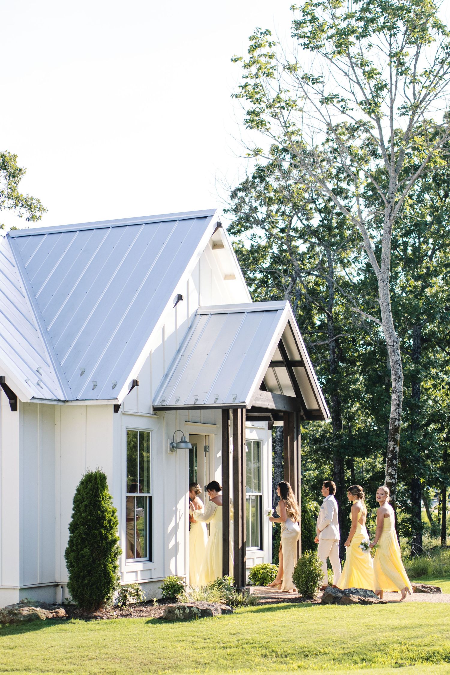 Bridesmaids in yellow dresses gather outside white chapel buildings.
