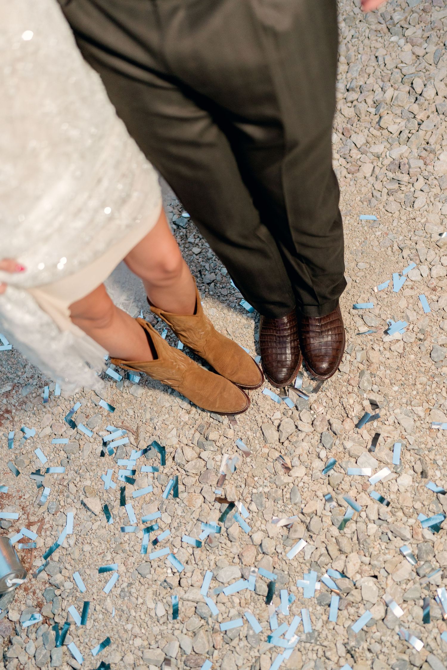 Close-up of bride in tan cowboy boots and groom in brown dress shoes standing on confetti-covered ground.