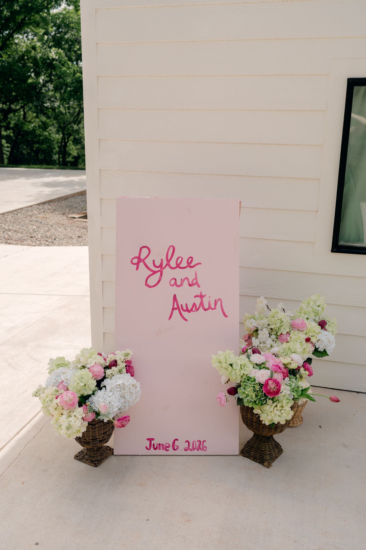 Pink wedding welcome sign for Rylee and Austin dated June 6, 2026, flanked by floral arrangements with pink and white blooms.