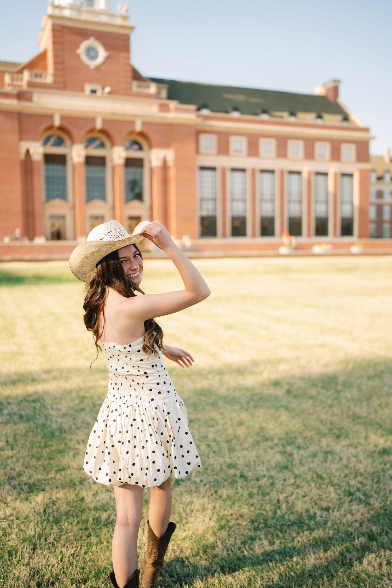 Person in polka dot dress and cowboy boots dances playfully in front of brick building.