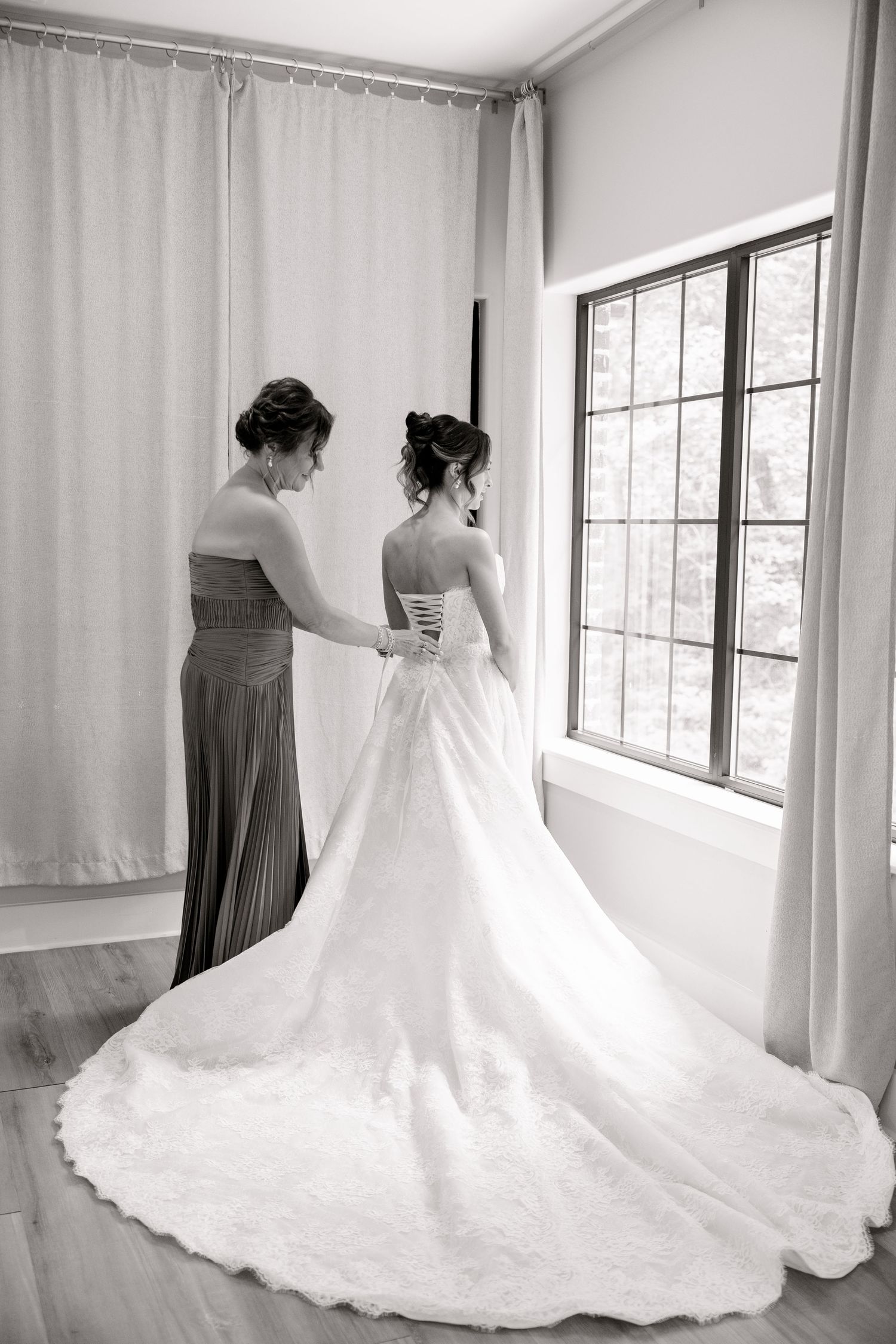 Black and white photo of a bride in a lace wedding gown being helped by a bridesmaid near a bright window.