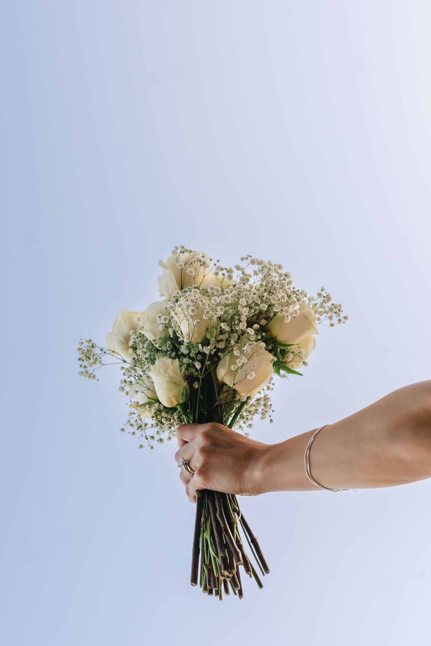 Hands holding a delicate bouquet of white roses and baby's breath against a light blue background.