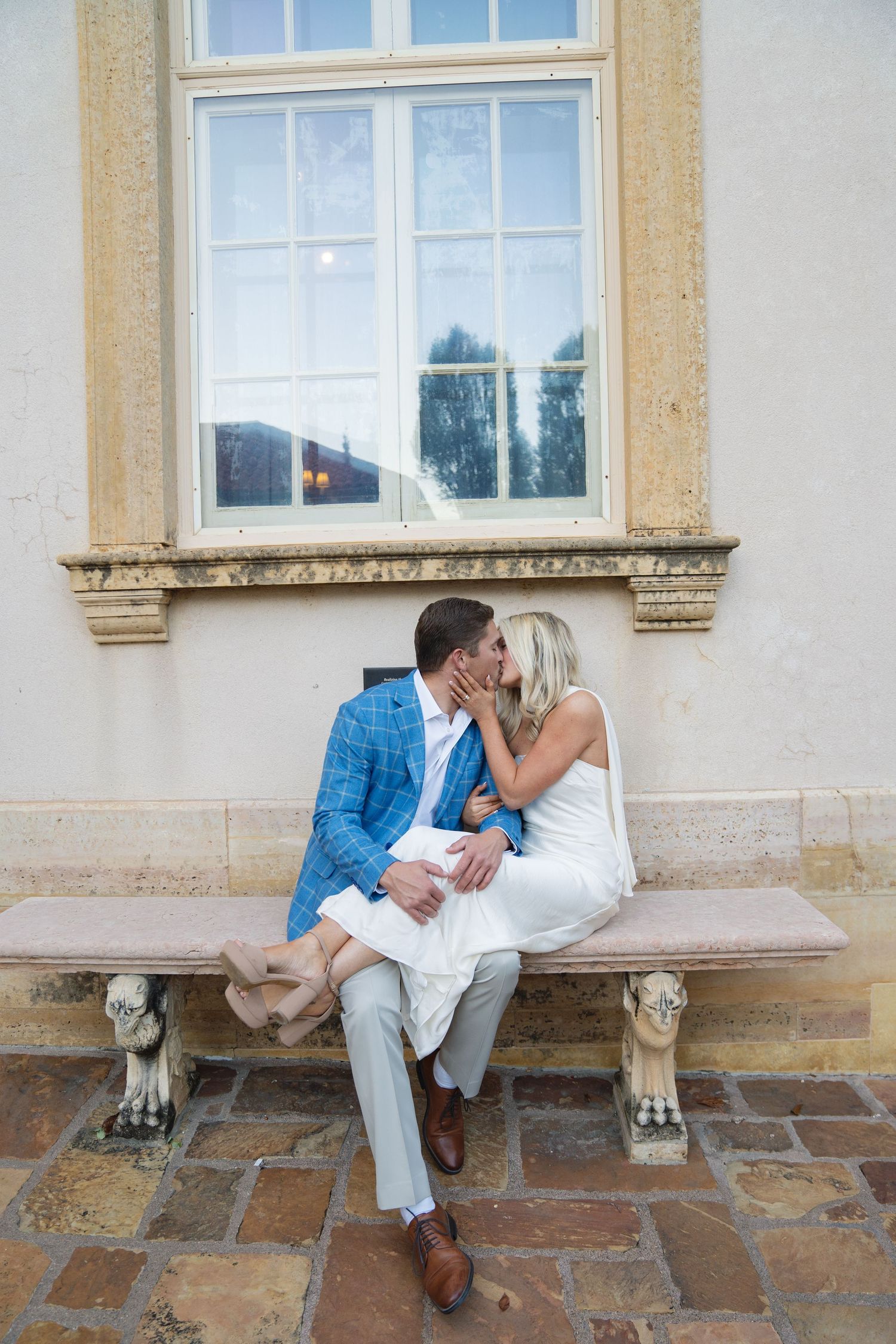 A couple shares a romantic moment on a stone bench by historic windows.