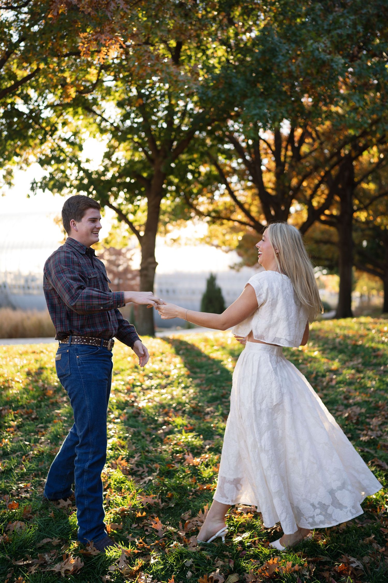 A couple dances together on grass with fall trees and golden sunlight in the background.