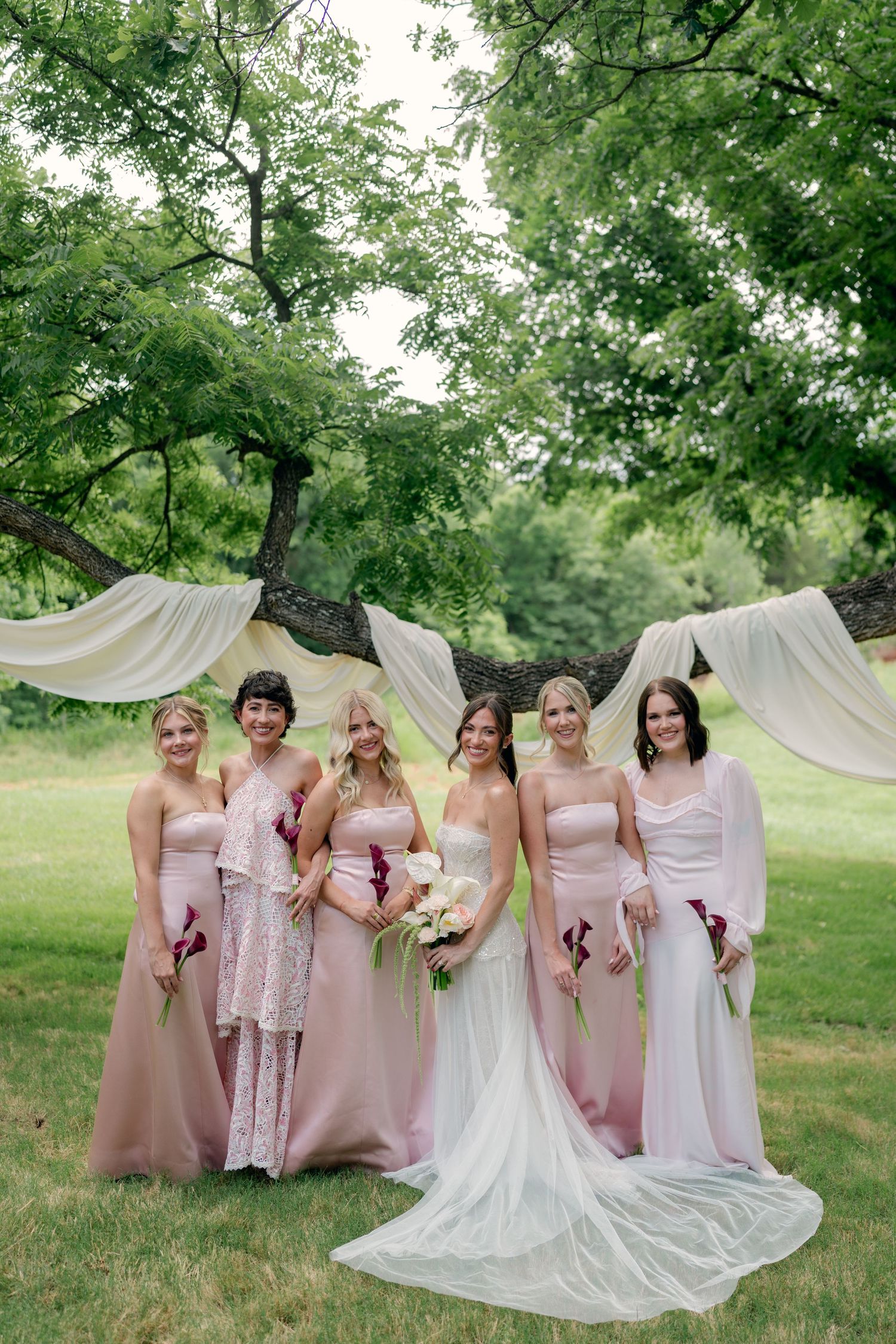 Bride and bridesmaids in pink dresses pose outdoors under a tree with white draping at a garden wedding.