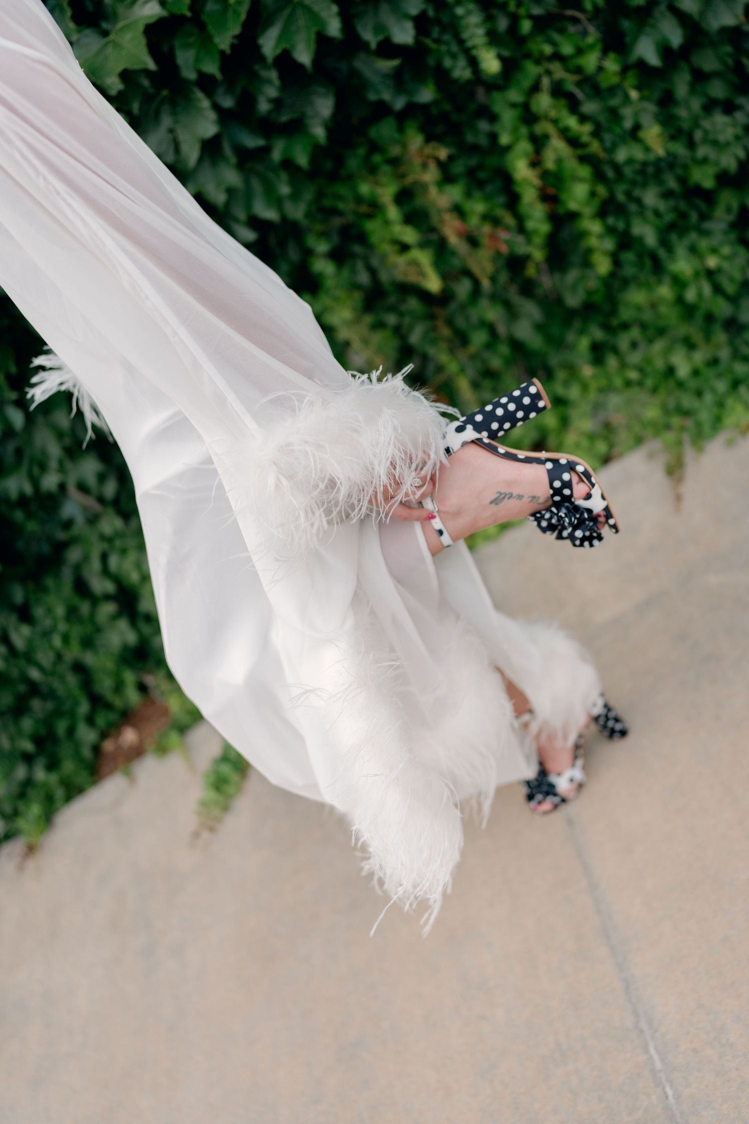 Close-up of bride wearing polka dot heels and feather-trimmed wedding gown hem against ivy wall backdrop.
