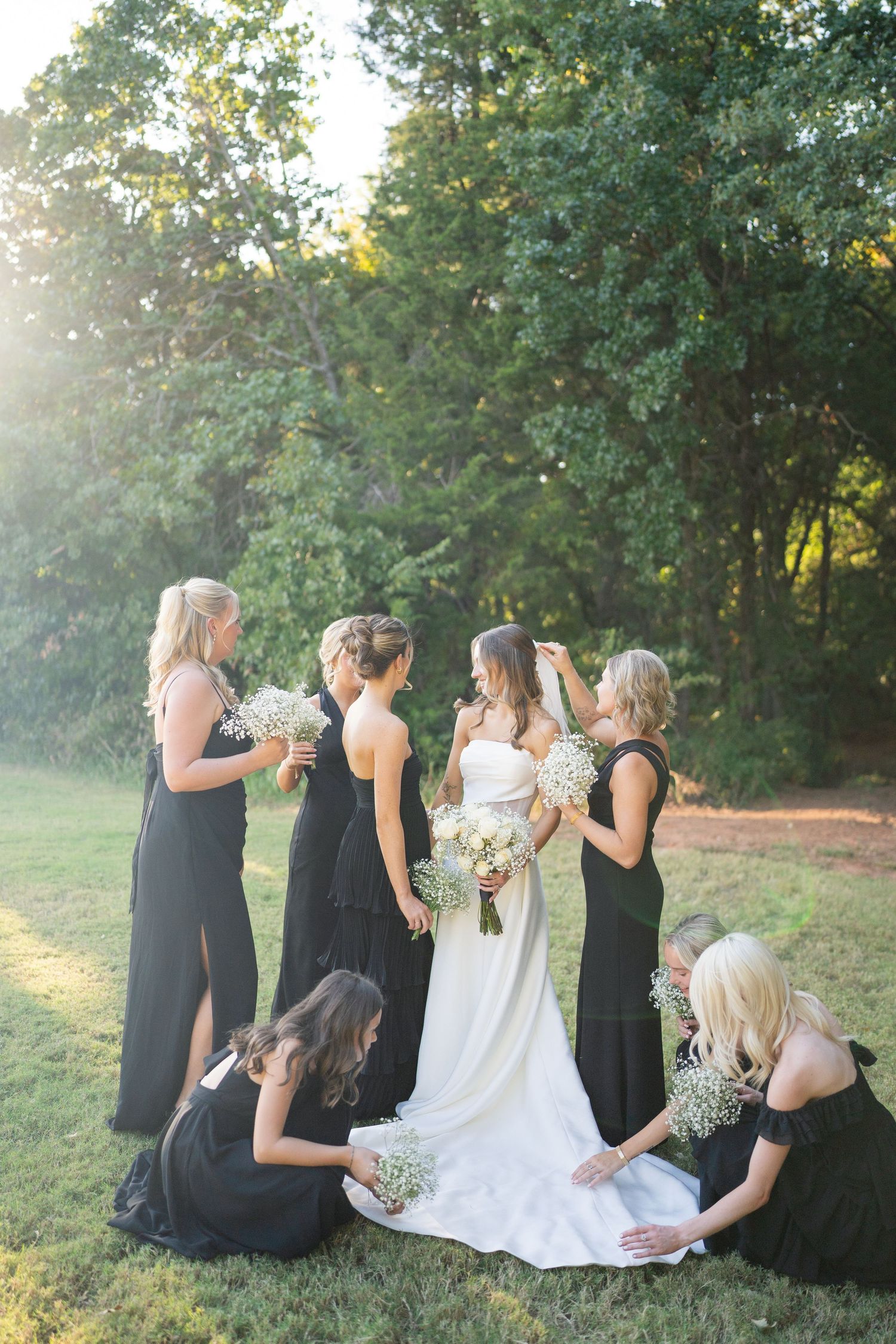 Wedding party in black dresses sitting together on grass during an outdoor celebration.