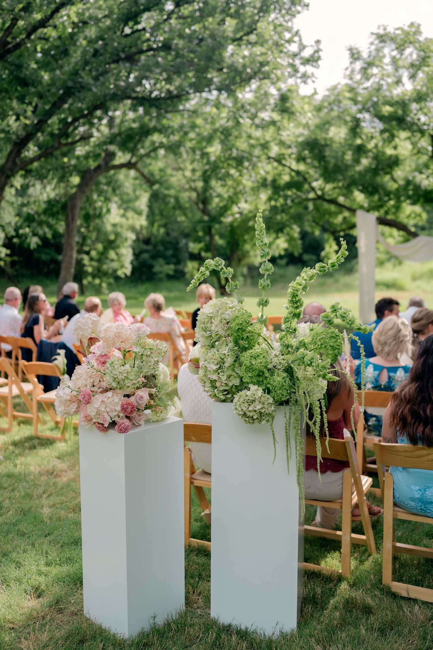 White pedestals with lush green and pink floral arrangements line the aisle of an outdoor wedding ceremony.
