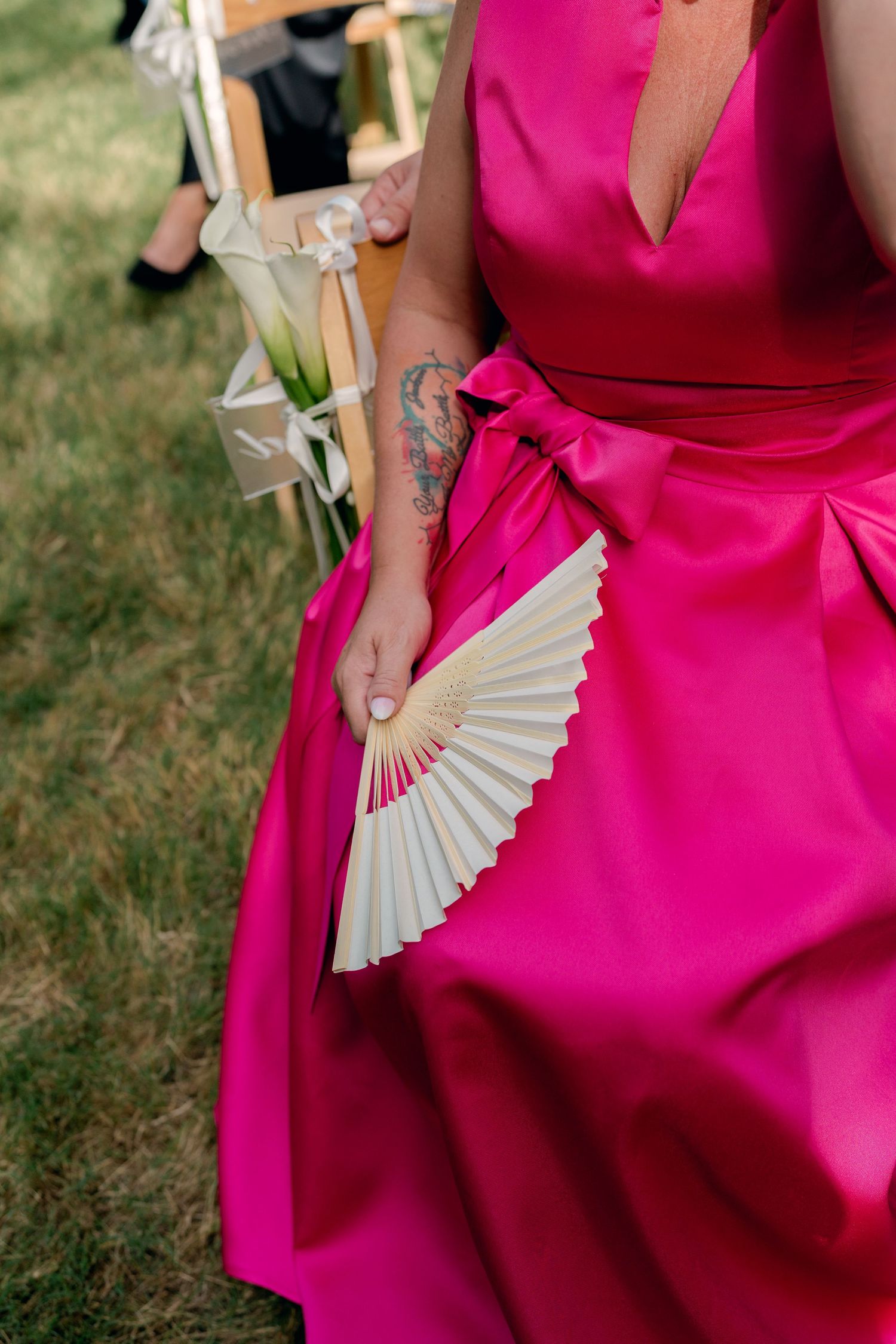 Wedding guest in vibrant fuchsia satin gown holding a white pleated hand fan at outdoor ceremony.