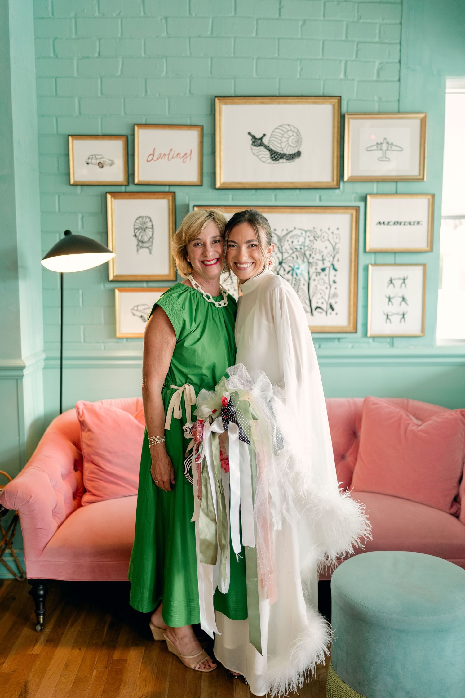 Bride in white feather-trimmed cape posing with woman in green dress against mint green wall with gallery art.
