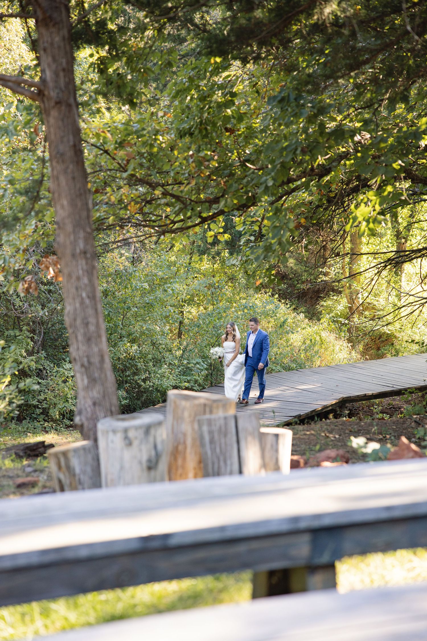 Wedding couple walking together on a wooden bridge surrounded by lush green trees in a park setting.