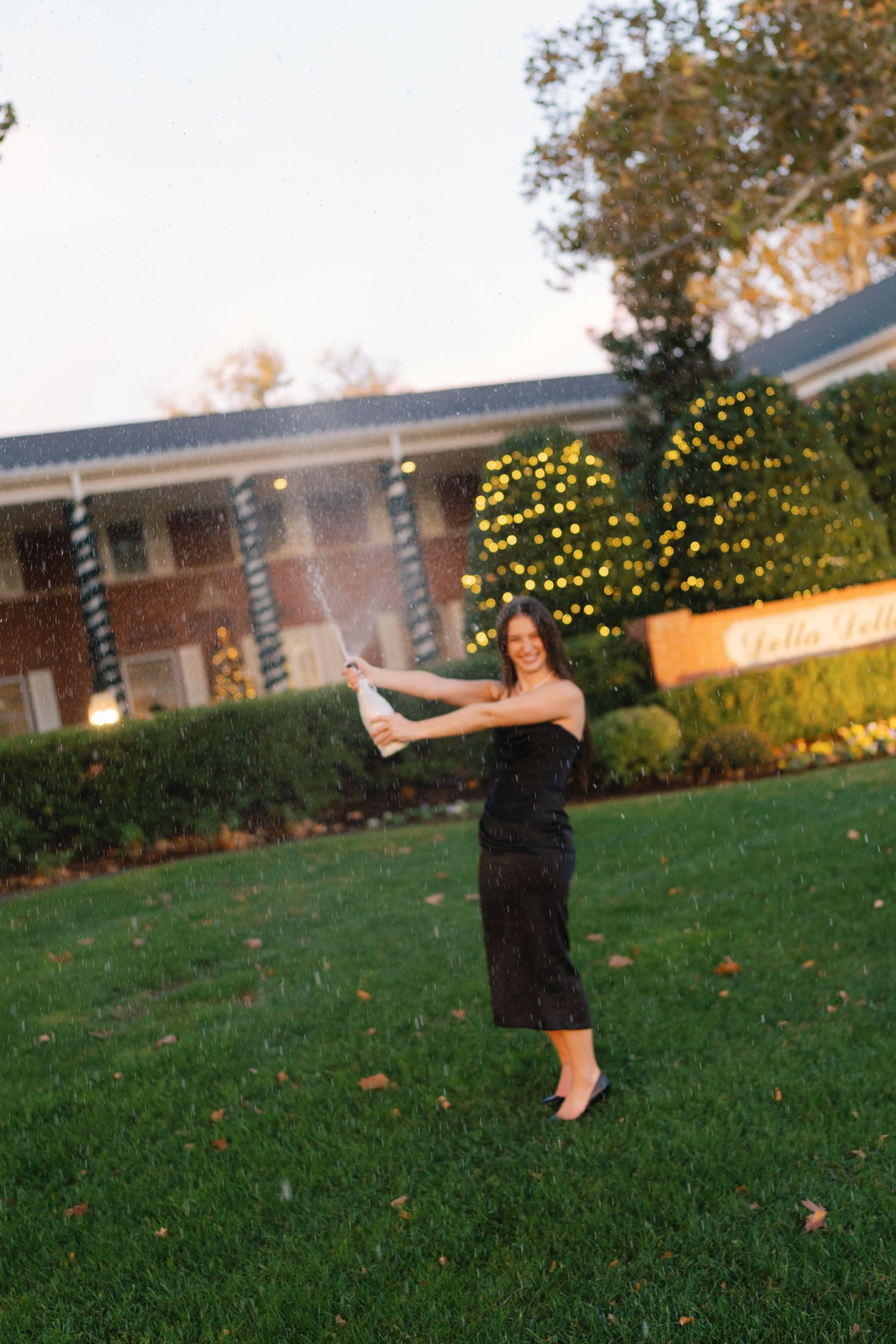 Someone in a black dress poses playfully on a lawn with holiday lights and brick buildings in the background at dusk.