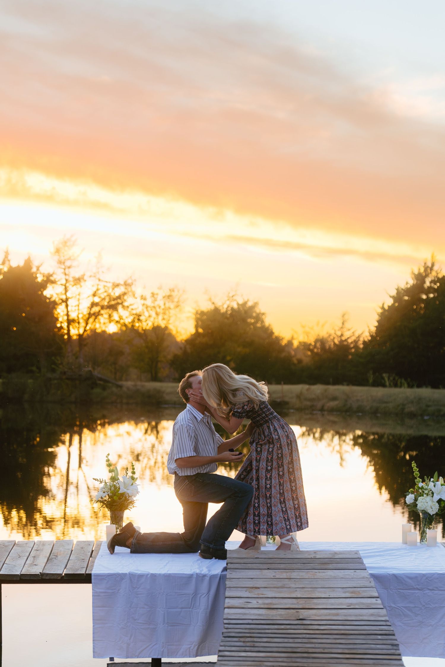 A romantic proposal scene on a wooden dock at sunset with a lake reflection in the background.