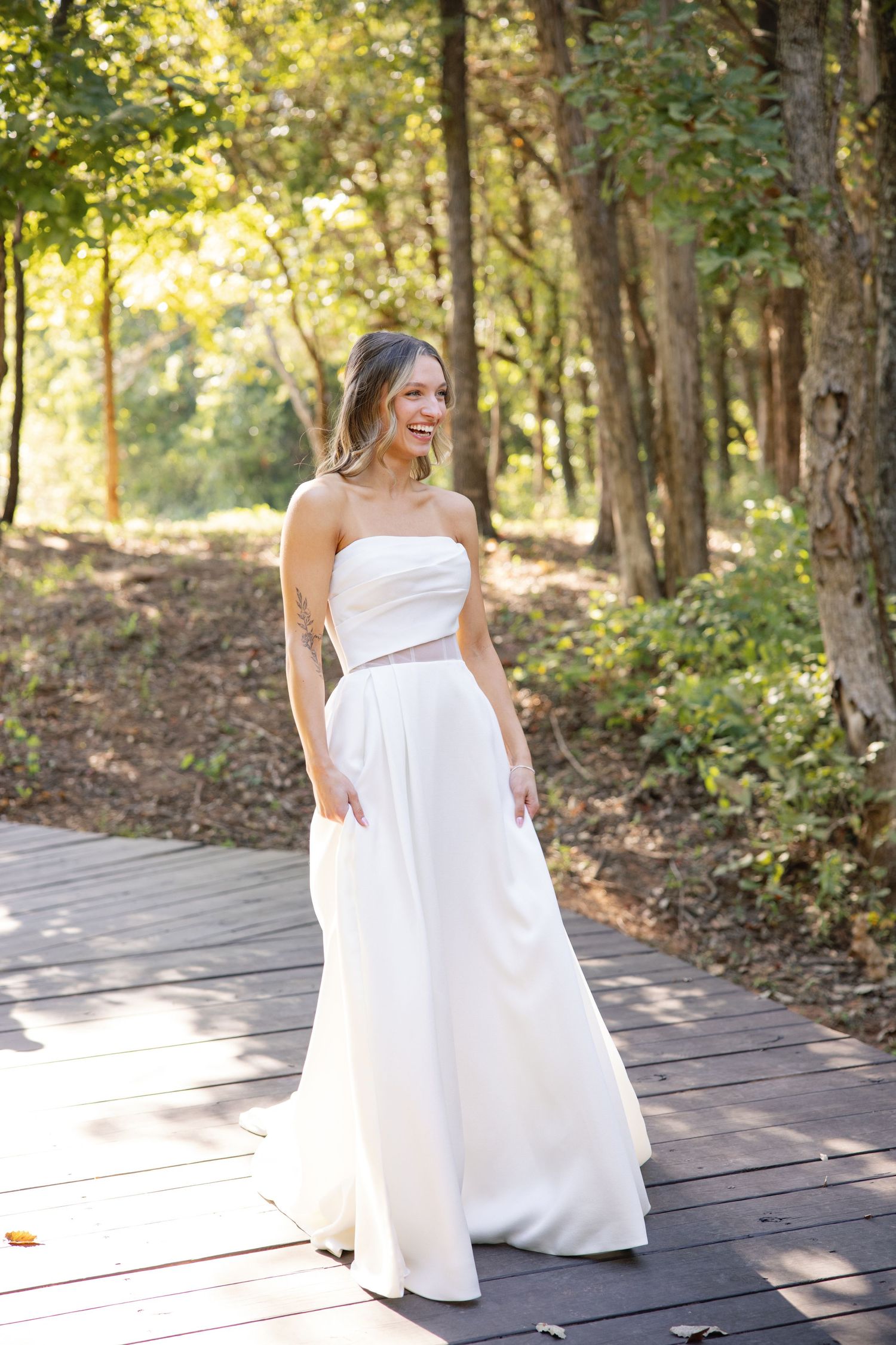 Modern bride in strapless white wedding gown poses on wooden walkway in sunlit forest.