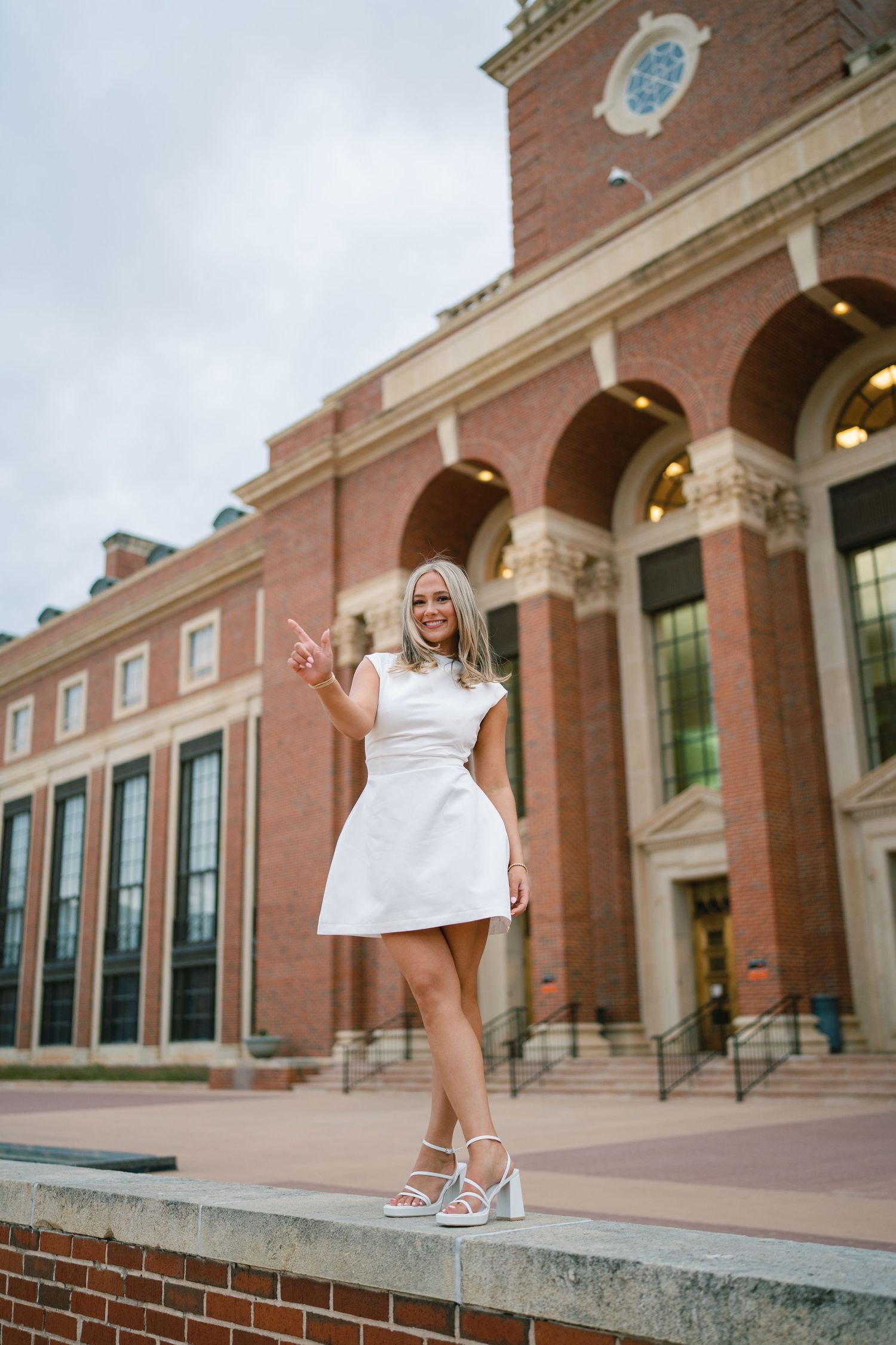 Person in white dress posing in front of grand brick building with arched windows.